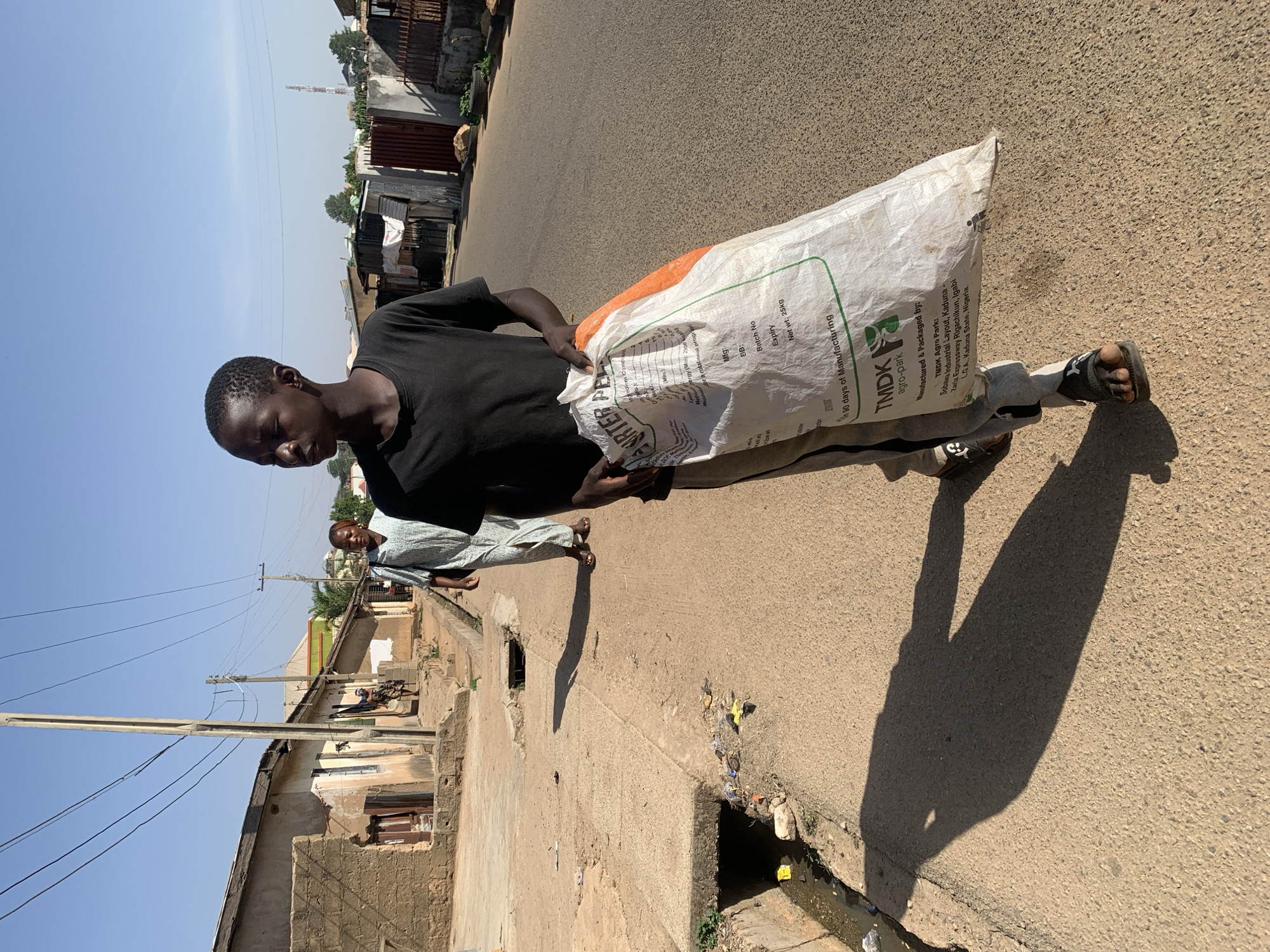 A young man carries a large, white sack with orange trim and printed text, possibly containing an agricultural product from TMDK Agro Park in Kaduna State, Nigeria, down a street in Jos, Nigeria. The environment consists of buildings, power lines, a concrete walkway, and a street with a drainage ditch. Other people are also visible in the background.
