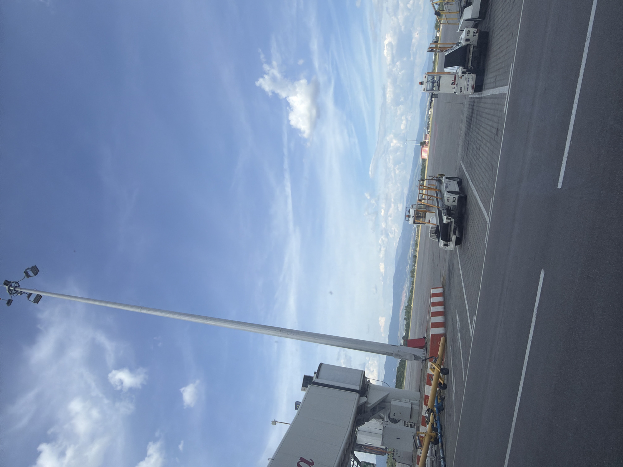 The image depicts an outdoor scene at an airport with a clear blue sky with scattered clouds. In the foreground, there is an asphalt surface with white lane markings. To the left, a jet bridge, identified by the partial letter "a" on its side, extends from a terminal building. A tall, silver light pole with multiple lamps is positioned near the jet bridge.
Further into the scene, on the tarmac, are several ground support vehicles. One appears to be a tow tractor, and another is a baggage conveyor belt. These vehicles are white and have markings like "TLC" and "GH 45108A" visible. In the background, there are red and white striped concrete barriers, and beyond that, a treeline and mountains under a hazy sky.
This scene is consistent with the area around the Sofia Airport in Sofia, Bulgaria, specifically near a gate where aircraft would dock. There are no people visible in the image. The environment suggests a sunny day at the airport, with the ground support equipment positioned for aircraft operations.