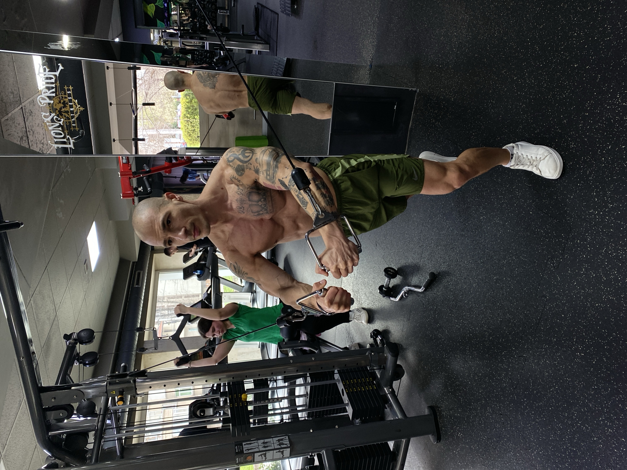 The image depicts a bald, shirtless man with tattoos performing a chest exercise on a cable machine in a gym. He is wearing green shorts and white sneakers. Another person in a green shirt is seen using a similar cable machine in the background. The gym has a mirror with the logo "Lion's Pride Fitness" reflected on it. The location is likely Pleven, Bulgaria, based on the gym's name and potential context. The gym's interior includes black rubber flooring with light-colored speckles, tiled walls, and weight training equipment.