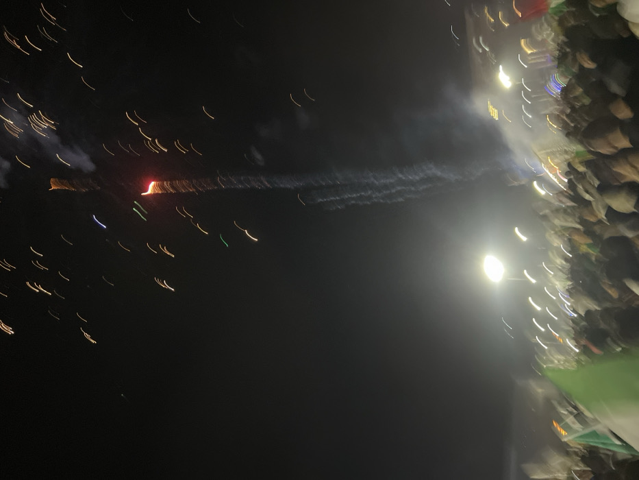 The image shows a nighttime scene with streaks of light, suggesting movement. On the left, a reddish point of light is surrounded by yellow streaks. A wisp of smoke trails away from it. On the right, a blurry illuminated area is visible, marked by vertical streaks of white and other colors, possibly indicating a crowd. The background is predominantly dark, and the overall impression is one of an event or display occurring at night.