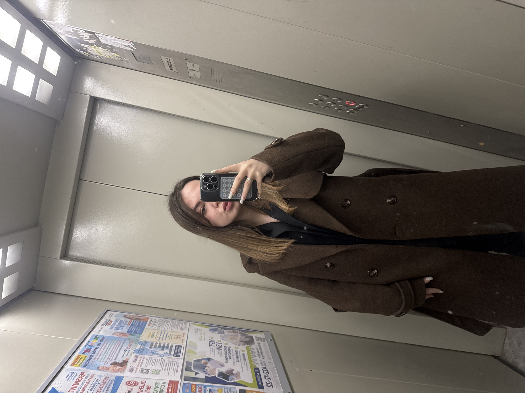 A woman in a brown overcoat is taking a selfie in an elevator. Elevator advertisements are visible on the wall, with the phone number 455-15-55, and "Orekhovo-Zuyevo." The elevator control panel is visible above the woman.