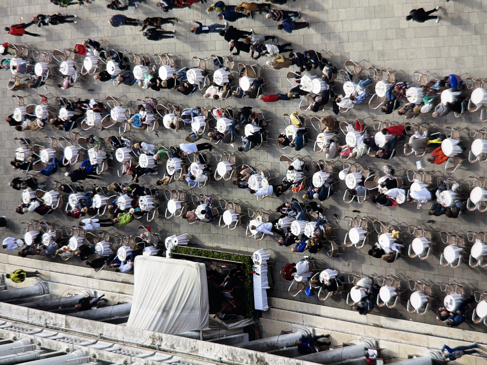 The image depicts an aerial view of a bustling outdoor cafe scene in a European city, possibly Venice due to the architecture and canal-like water features suggested by the context. The cafe is filled with numerous small, round tables, each covered with a white tablecloth and surrounded by chairs. Many patrons are seated at these tables, engaged in dining, drinking, and conversation. The scene includes a variety of individuals, men and women, dressed in casual attire. Some people are actively interacting, such as ordering food, gesturing, or looking at each other, while others appear to be relaxing or observing their surroundings. In the background, there are stone architectural elements, including columns and arches, suggesting an historic urban setting. A street musician or performers can be seen playing instruments in one section of the cafe, adding to the ambiance. The ground is paved with stone tiles, and there is some greenery integrated into the cafe's design. The overall atmosphere is one of lively social activity and enjoyment of a pleasant outdoor environment. The image does not show any specific landmarks or indications that would place it in Kyiv, Ukraine.