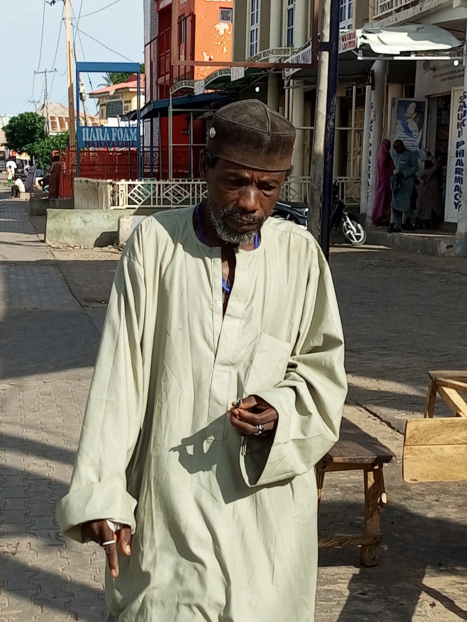 The media file shows a medium shot of an elderly Black man walking on a paved street in what appears to be a market or commercial area in Arawa, Nigeria. He is wearing a light-colored, long-sleeved robe and a dark cap. His face is weathered, and he has a gray beard and mustache. He is holding a small object in his right hand, and his left hand is resting on his chest, holding another object.
The background suggests a bustling street with several buildings. To the left, there's a building with orange and white facades and a blue sign that reads "HARA FOAM". Further down the street, there are shops with awnings and signs, including one that appears to say "SAUKKI PHARMACY". Several other people are visible in the background, some walking and others standing, suggesting it is daytime, likely late morning or early afternoon, given the strong shadows cast on the ground. The weather appears to be sunny and warm. The man's expression is neutral, and the overall scene conveys a sense of everyday life in a Nigerian town. There are also wooden benches and tables visible on the side of the street.