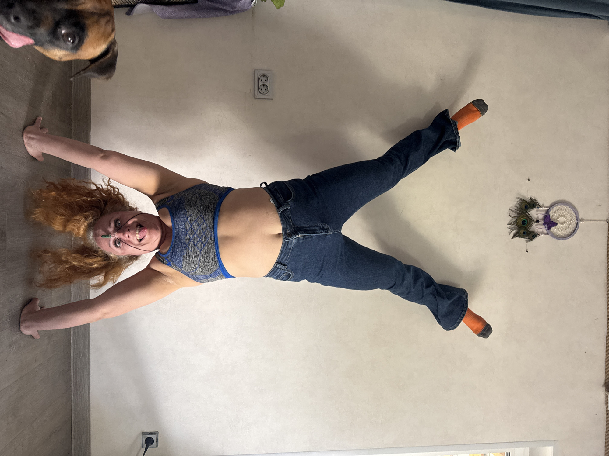 A woman is lying on her back on a beige floor and white wall in what appears to be a room in a building, possibly in the city of Chaikovsky, Russia. She is wearing a gray and blue sports bra, blue jeans, and orange socks. Her arms are outstretched to her sides, with her hands on the floor. Her legs are spread apart. She has red hair and is sticking out her tongue. A dog is visible in the upper left corner of the image. An electrical outlet is on the wall behind her. A dream catcher hangs near the top right corner of the wall. Another outlet is behind the woman's hair. A window is at the bottom of the image.