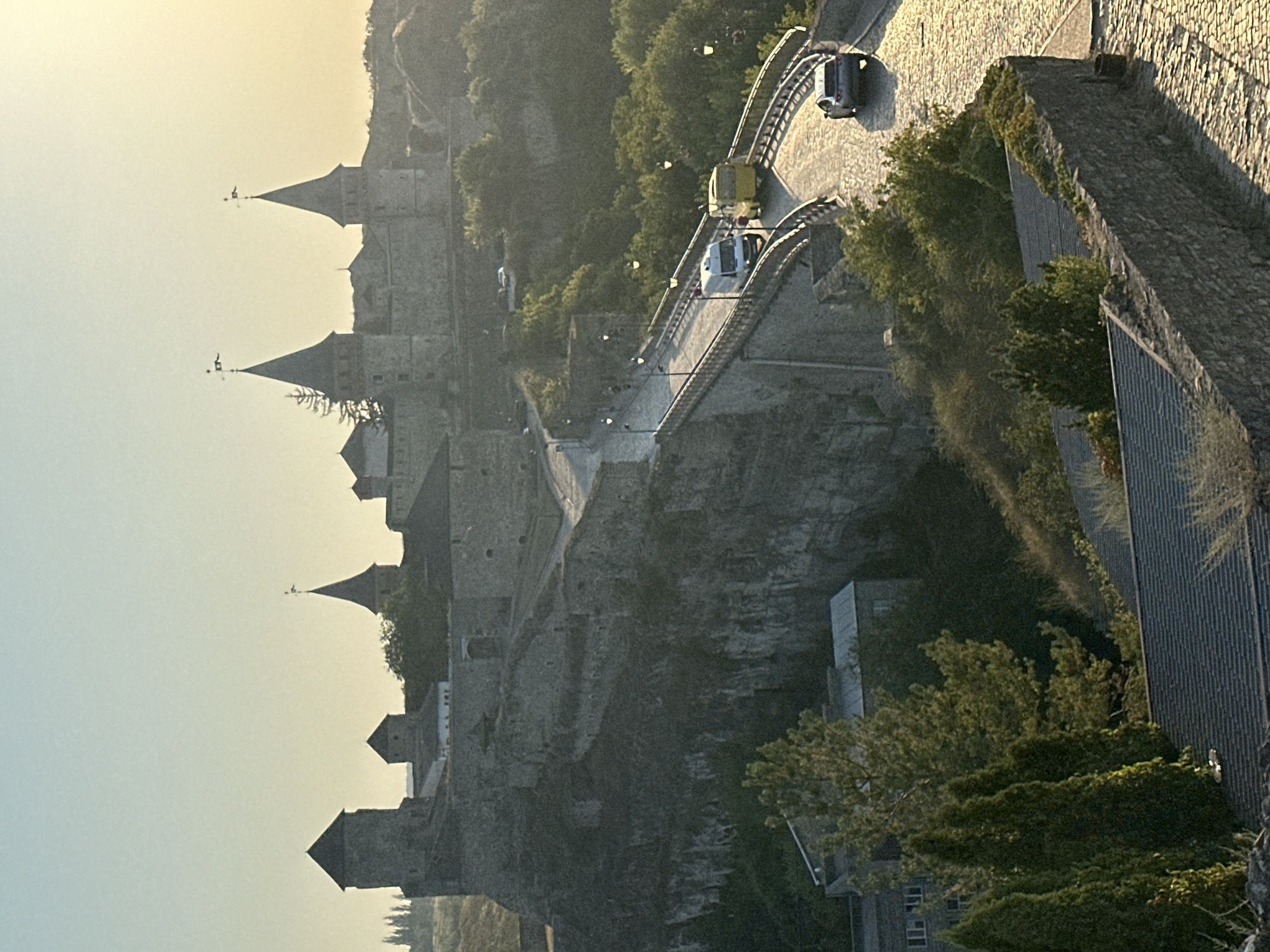 The image shows the Kamianets-Podilskyi Castle in Kamianets-Podilskyi, Ukraine, bathed in the warm light of a sunset or sunrise. The imposing stone fortress, with its numerous towers and battlements, is perched atop a rocky cliff. A stone bridge, paved with cobblestones, leads towards the castle, with a white van, a yellow bus, and a silver car traversing it. The bridge is lined with lampposts that cast pools of light. Lush green trees and vegetation surround the castle and the bridge, adding a natural element to the scene. In the foreground, more stone walls and rooftops of nearby buildings are visible, some covered in ivy. The sky above is a soft gradient of blue and yellow, indicating the time of day.