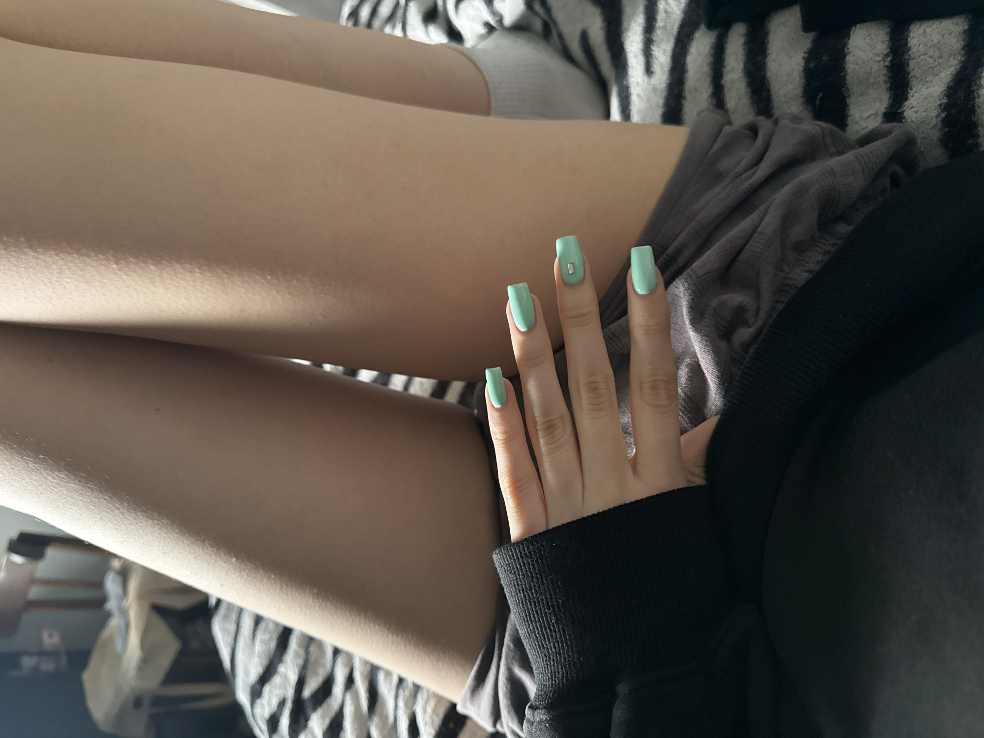A person's legs are crossed, with a hand resting on the upper leg. The fingers are adorned with mint green nail polish. The person is wearing grey shorts and a black long-sleeved shirt. The background features a zebra-striped fabric. The location is not discernible.