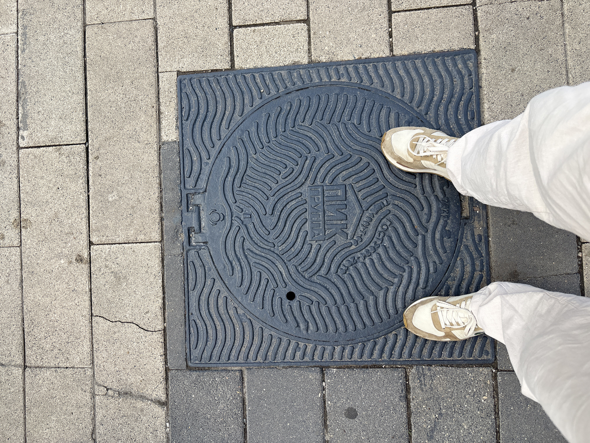 A ground-level perspective shows a person standing on a square metal manhole cover set into a paved surface. The manhole cover is dark gray and features a circular design with repeating wavy lines and Cyrillic text near the center. The paved surface around the manhole cover consists of gray rectangular blocks. The person is wearing off-white or beige sneakers and white pants. The scene appears to be outdoors, likely in a city or urban environment. No specific location is identified.