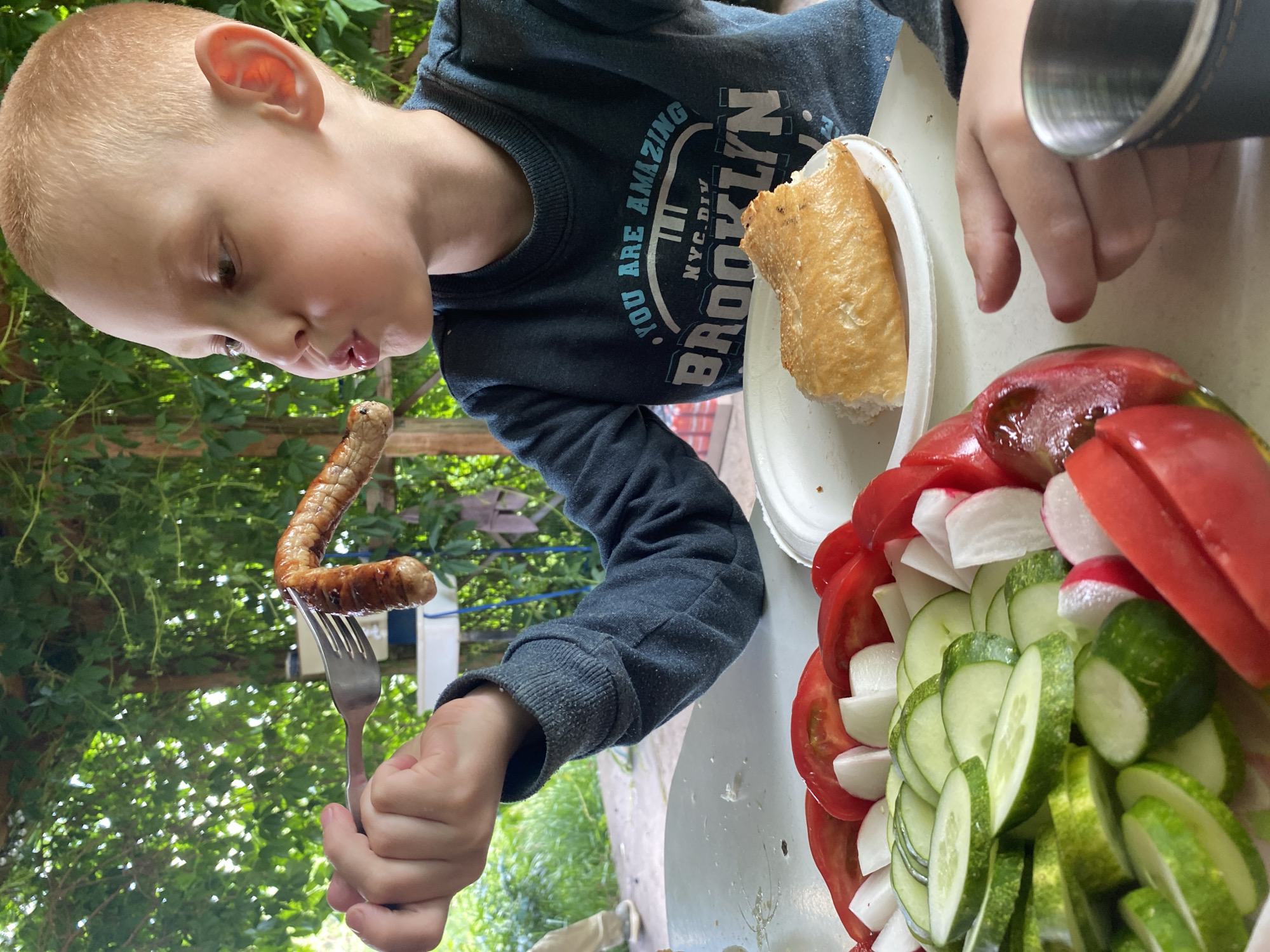 A close-up, high-angle shot captures a young boy during an outdoor meal. The boy, with short, light-colored hair and fair skin, is seen in profile, looking intently downwards at a grilled sausage or piece of meat bent into a 'U' shape on a fork held in his right hand. His lips are slightly pursed, conveying focus or anticipation. He is wearing a dark blue long-sleeved sweatshirt that displays the text "YOU ARE AMAZING" along with "NYC.DIV" and "BROOKLYN" written vertically.
On the table in front of him, to his right, is a white paper plate holding a piece of bread or a bun. Further to the right, a colorful salad is arranged in a bowl, featuring neatly sliced red tomatoes, white radishes, and green cucumbers. A portion of an adult's hand and a dark metal cup or thermos are visible on the far right side of the frame.
The background is filled with lush green foliage, indicating a sunny daytime outdoor setting, likely a garden or patio. The scene portrays a casual, relaxed mealtime, consistent with the provided location context of Chaikovsky, Russia.