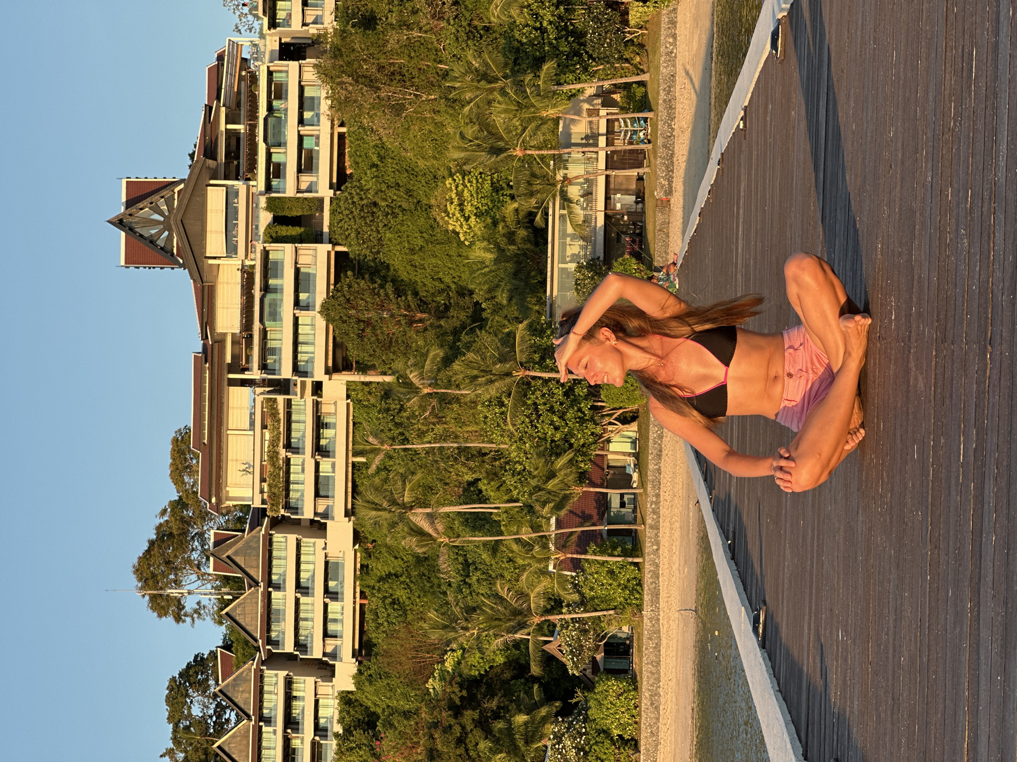 A young woman with long brown hair, wearing a black bikini top with pink trim and pink shorts, sits in a lotus pose on a wooden pier. She is facing towards the right of the frame, with her right hand touching her forehead and her head tilted back. The pier extends forward towards the water, with the ocean on the left and a sandy beach on the right. Behind the beach, lush green vegetation covers a hillside, topped by a large, multi-story building with a distinctive architectural style, likely a resort. The building features multiple balconies, large windows, and a roof with traditional Thai design elements. Palm trees are interspersed among the vegetation and near the beach. The scene is illuminated by the warm, golden light of a setting or rising sun. The location is Baan Taling Ngam, Thailand.