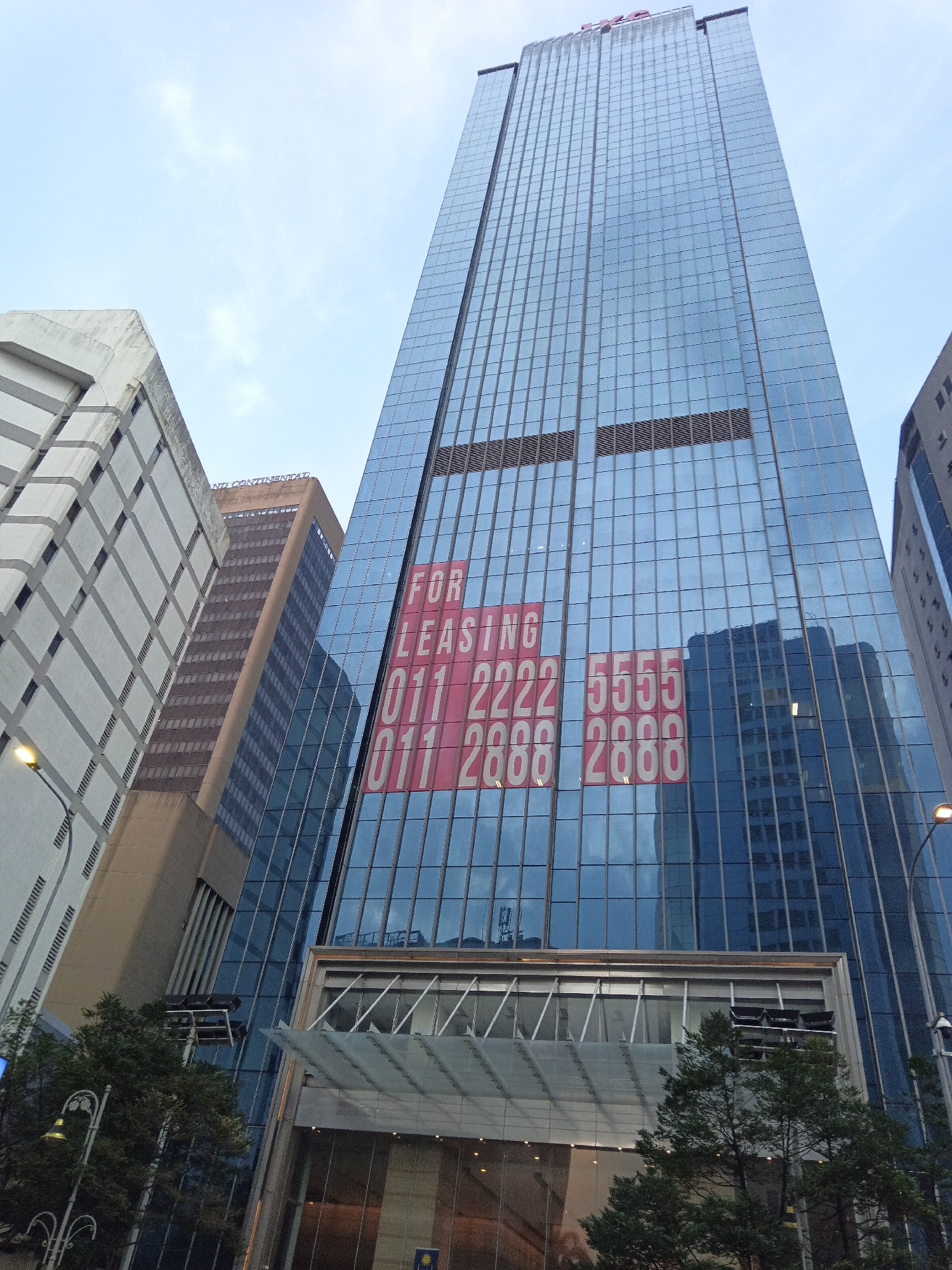 The image is an eye-level, low-angle shot of the exterior of a modern, glass-fronted skyscraper in Kuala Lumpur, Malaysia. The skyscraper is characterized by its numerous reflective blue glass panes, which create a mosaic of building reflections. Prominently displayed on the glass facade are large, red and white signs indicating "FOR LEASING" along with phone numbers: "011 2222 5555" and "011 2888 2888". The sky above is overcast, casting a diffused light on the scene. To the left of the main skyscraper, another building with a more textured, grey and white facade is visible, featuring rows of recessed windows. Further to the left and in the mid-ground, a brown, rectangular skyscraper is also present, showing a different architectural style. The foreground of the image shows the top of trees and a streetlamp with a decorative pole, suggesting an urban streetscape. The overall impression is one of a bustling commercial district.