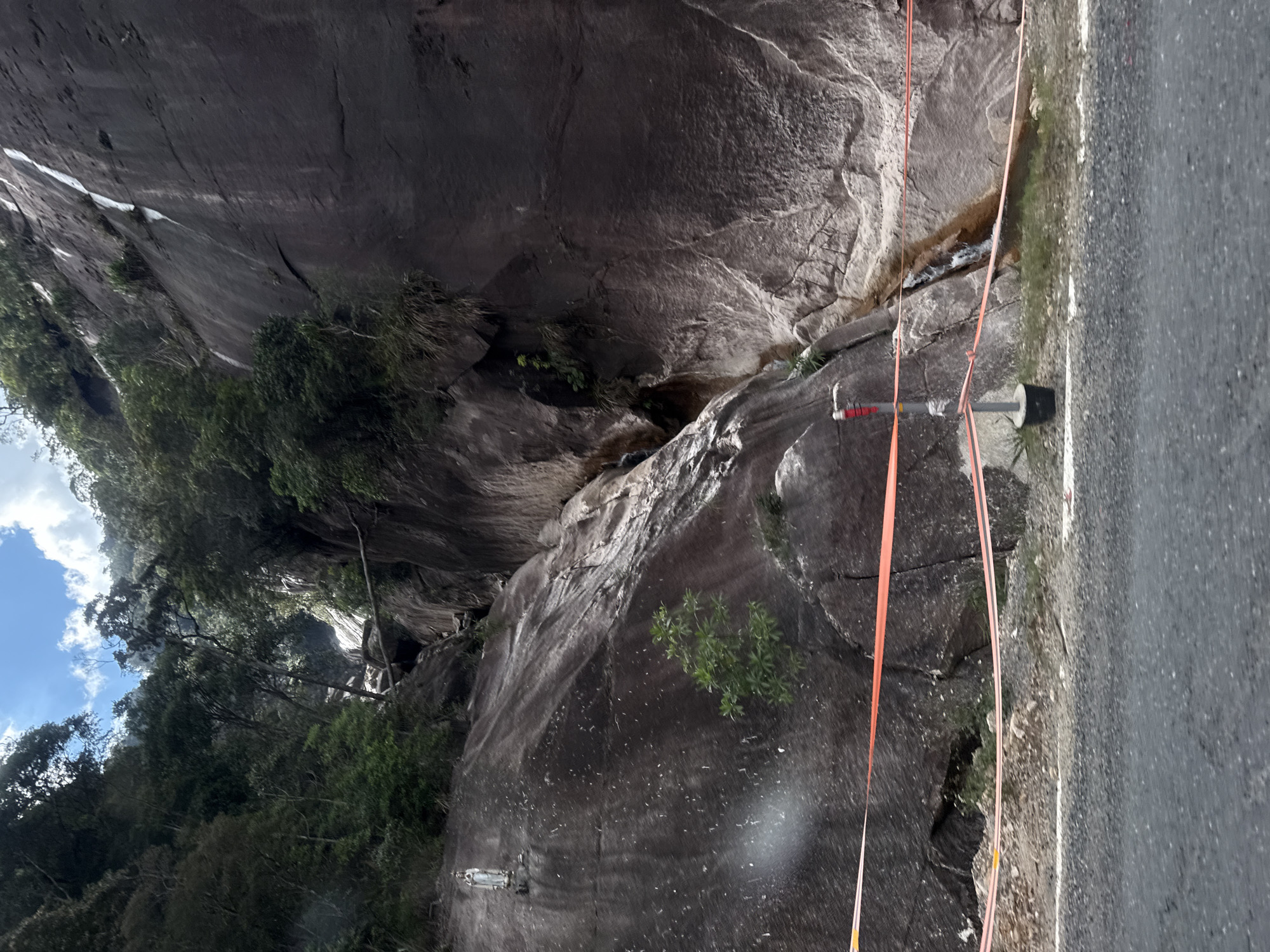 The image depicts a rocky cliff face adjacent to a paved road. The cliff is dark gray and appears to be made of granite, with visible cracks and fissures. Patches of green vegetation, including small trees and bushes, grow out of the rock in several places. A narrow stream of water trickles down the cliff face in a central gully.
Two orange ropes are stretched diagonally across the scene, one appearing to be secured to a metal pole with a black cylindrical object attached. This suggests some form of activity or construction is taking place, possibly related to rock stabilization or a survey. The ropes are taut and extend from the cliff face towards the road.
The road itself is asphalt and shows a white dividing line. The surrounding area is a natural landscape, with lush green trees visible in the upper left portion of the image, and a cloudy blue sky. The lighting suggests it is daytime, with diffused sunlight. There is no clear indication of the specific time of day or weather conditions beyond the clouds.
The location is identified as Nam Khanh Vinh, Vietnam. There is no visible text in the image. The scene conveys a sense of rugged natural beauty combined with human intervention.