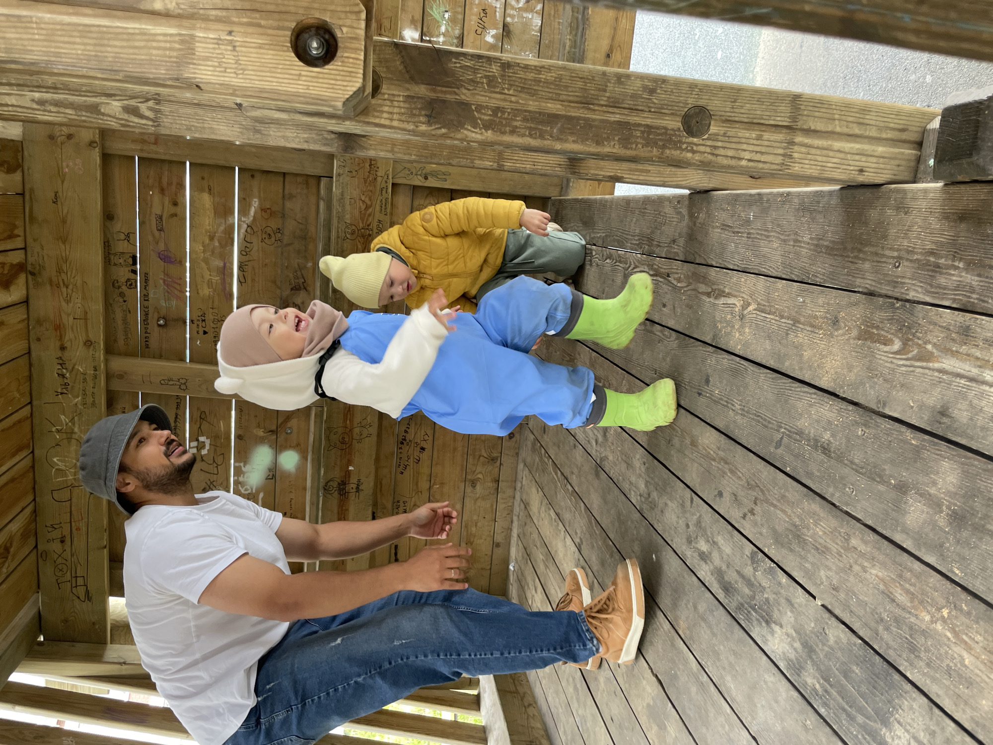 This media file captures a playful moment between a man and two young children inside a rustic wooden structure, likely an outdoor play area or pavilion in Izhevsk, Russia.
The man, positioned on the left, wears a white t-shirt, blue jeans, brown boots, and a grey bucket hat. He has a beard and mustache and is looking upwards with a slight smile. In the center, a young child, possibly a toddler, is dressed in a light blue full-body suit, a light beige balaclava-style hat, and bright green rubber boots. This child is leaning back or lying down, mouth open in an expression of joy or laughter, looking towards the top of the frame. Further back and to the right, another young child wears a yellow puffy jacket, green pants, and a light yellow beanie, observing the scene while leaning against the wooden wall.
The setting is characterized by unpainted wooden planks forming the walls and floor, typical of a simple, sturdy outdoor construction. The walls are covered extensively with graffiti, including various scribbles, drawings, and text in Cyrillic. Notable visible text includes "ищю," "Ульяна," "сука," and "рикв раБлоксе KIL MAYZZ," along with several phone numbers or numerical sequences like "895084083" and "99010201." The children's warm clothing suggests it might be a cool or damp day, possibly spring or autumn. The overall atmosphere is light-hearted and joyful, capturing a family interaction in a casual outdoor environment.