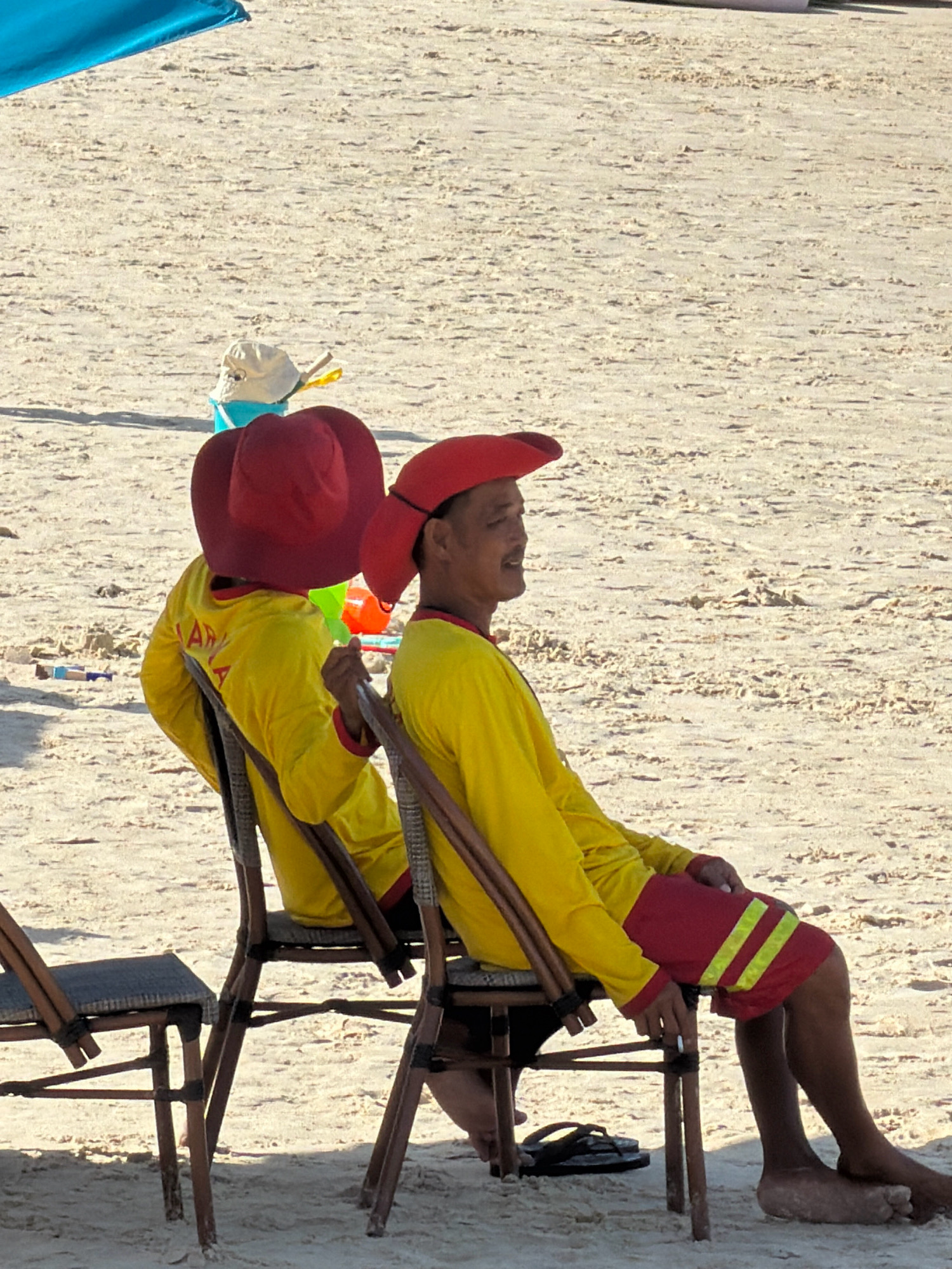 Two individuals wearing yellow long-sleeved shirts and red hats are seated on chairs on a sandy beach. The individual on the right is facing to the right, looking out towards the sea. They are wearing red shorts with two yellow reflective stripes. The individual on the left is facing away from the camera, with their back to the viewer. They are also wearing a yellow shirt and a red hat. In the background, there are beach toys, including a blue bucket with a white hat and a small shovel. The sand appears dry and lightly textured. A portion of a blue umbrella is visible in the upper left corner of the image. The scene suggests a relaxed atmosphere, possibly indicating a break for lifeguards on duty. The location is identified as Nha Trang, Vietnam.