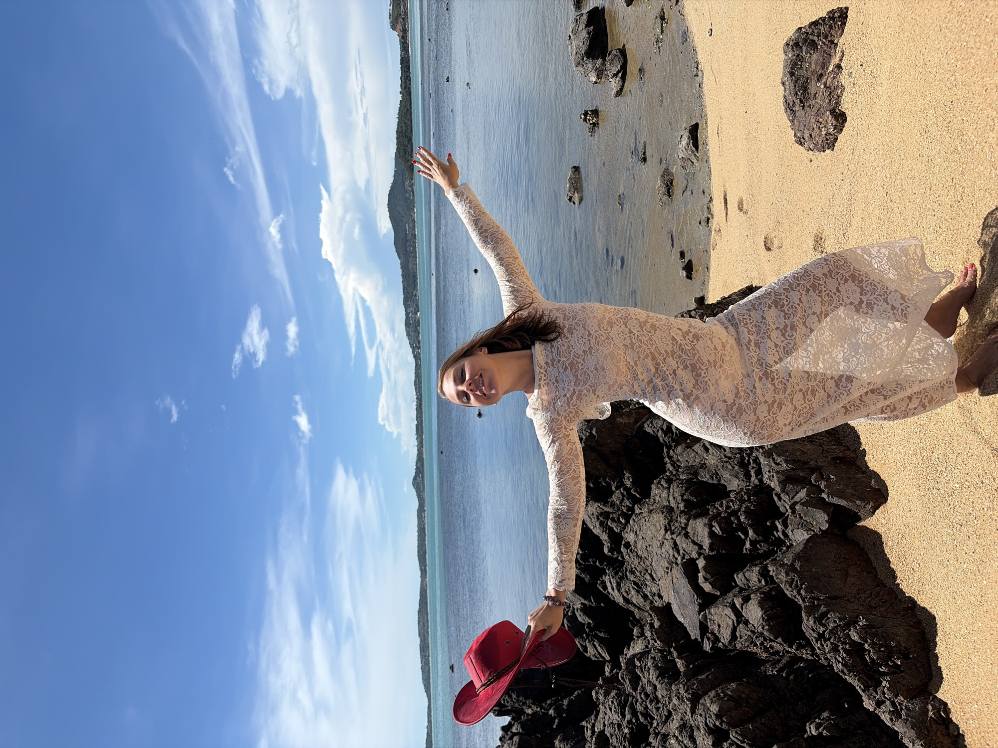 A woman stands on a sandy beach in Baan Bo Phut, Thailand, holding a red hat in her left hand. She is wearing a long, white lace dress and her right arm is extended towards the water. The ocean is calm and blue, with a distant island and green hills visible. The sky is bright blue with scattered white clouds. Dark volcanic rocks are on the left side of the image, and smaller rocks are scattered on the beach and in the water.