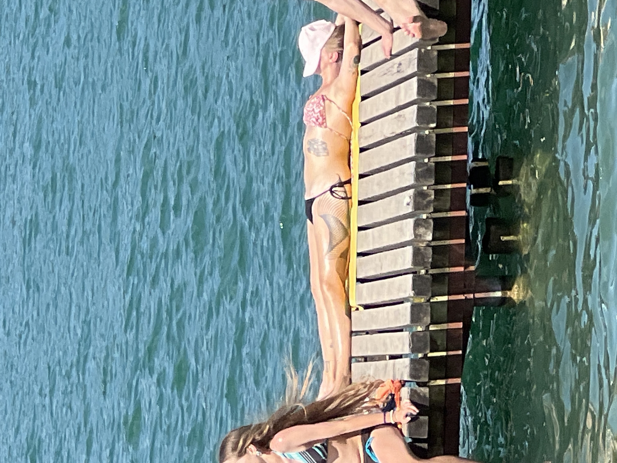 The image depicts a sunny day at a waterside location, possibly a pier or a dock, in Vilnius, Lithuania. In the foreground, a woman with long, light brown hair is sitting on the edge of a wooden pier, wearing a striped bikini top and dark bottoms. She is looking to her left. To her right, another person is lying on their stomach on a yellow mat on the pier, sunbathing. This person is wearing a pink and white patterned bikini and a light pink baseball cap. They have tattoos visible on their upper back and leg. Their arms are resting by their head. Partially visible on the far right is another person's legs and feet, also on the pier. The pier is constructed of wooden planks with dark supports visible underneath. The background is filled with shimmering blue water, indicative of a lake or a river. The overall scene suggests a leisurely summer activity.