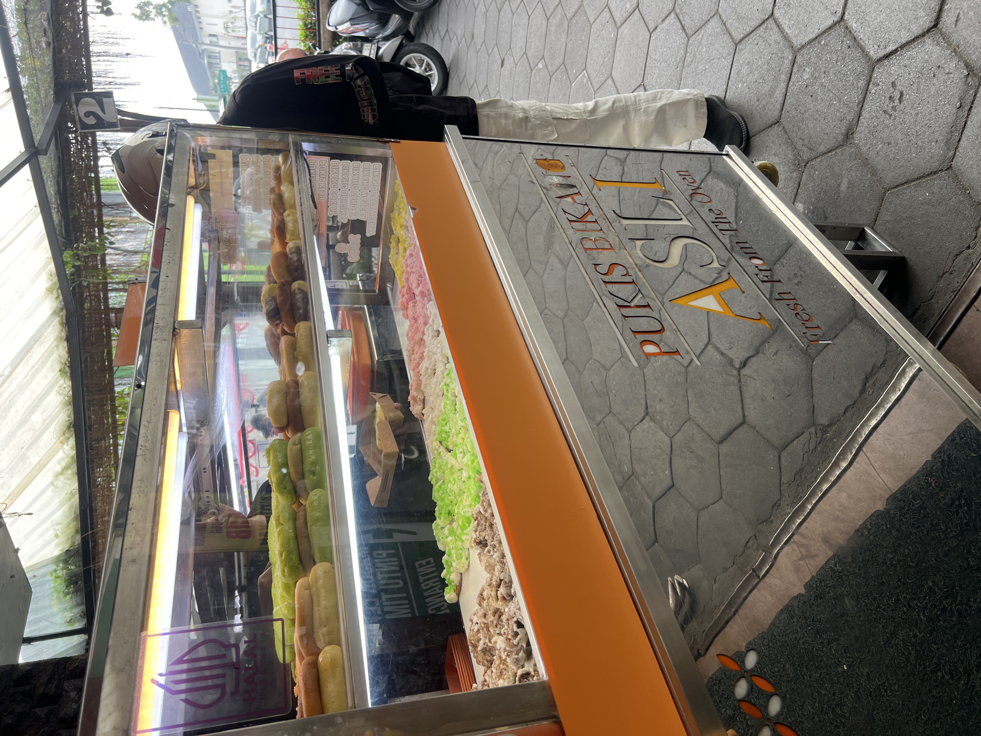 A mobile food cart, identified by the text "PUKIS BIKANG ASLI Fresh From The Oven" on its reflective surface, is parked on a hexagonal-tiled sidewalk. The cart features a refrigerated display case filled with colorful pastries, including what appear to be donuts and small cakes in various hues of green, pink, yellow, and white. Some pastries are coated in sugar or a similar granular topping. The cart is illuminated by bright fluorescent lights within the display. A person, partially obscured by a helmet and wearing a black long-sleeved shirt and light-colored cargo pants, stands behind the cart, attending to the food. The background shows a street scene with parked motorcycles and cars, suggesting an urban environment in Yogyakarta, Indonesia. The cart's exterior is a mix of polished metal and an orange frame.