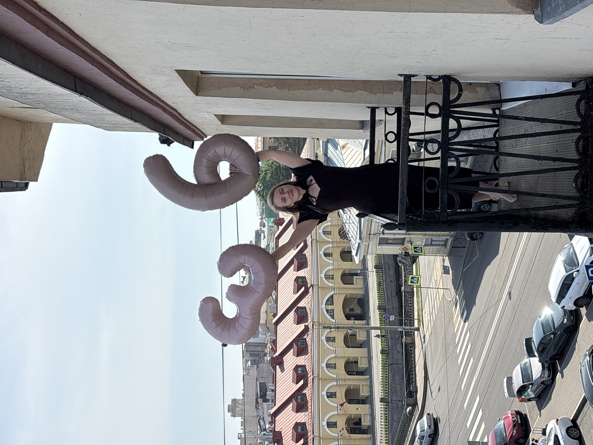 A woman stands on a balcony holding large foil balloons shaped like the numbers "3" and "9". She is wearing a black dress. The balcony has a black, ornate metal railing. Below the balcony is a street with a crosswalk and cars parked on either side. In the background are buildings with architectural details like arched windows. The street scene indicates a European city, specifically Saint Petersburg, Russia, suggested by the architecture and layout.