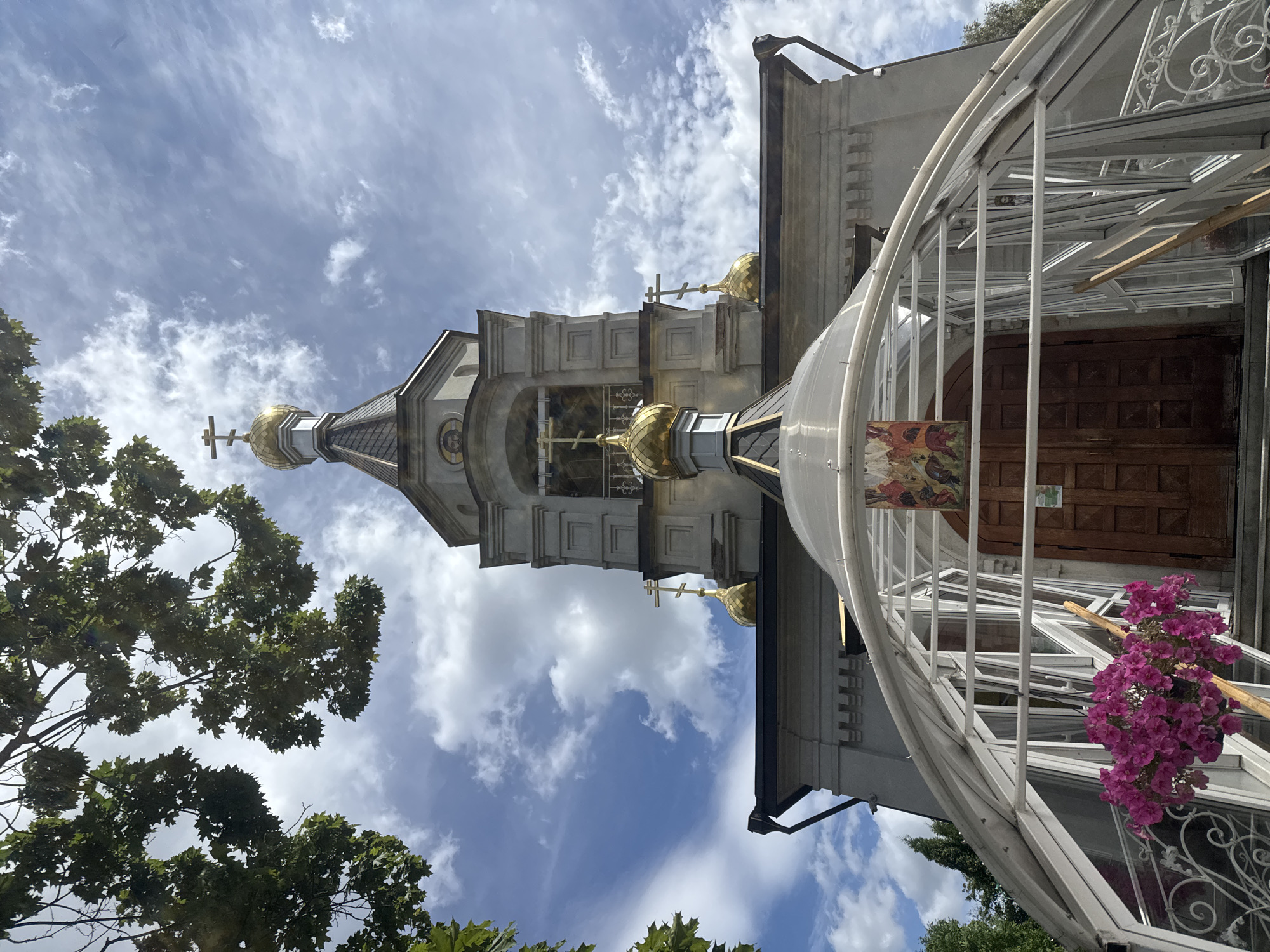The image shows the bell tower and entrance of an Orthodox church, likely in Sovkhoz imeni Lenina, Russia. The bell tower is a light gray structure with several levels, topped by a gilded dome with a cross. Below the bell tower, the main body of the church is visible, also in a light gray stone. The entrance is covered by a modern, arched greenhouse-like structure with white metal framing and clear panels. Inside this structure, a large wooden door is visible, above which hangs an icon. A cluster of vibrant pink flowers is in bloom in the foreground, in front of the greenhouse structure. The sky is blue with scattered white clouds, and the branches of a leafy tree extend into the upper left corner of the frame.