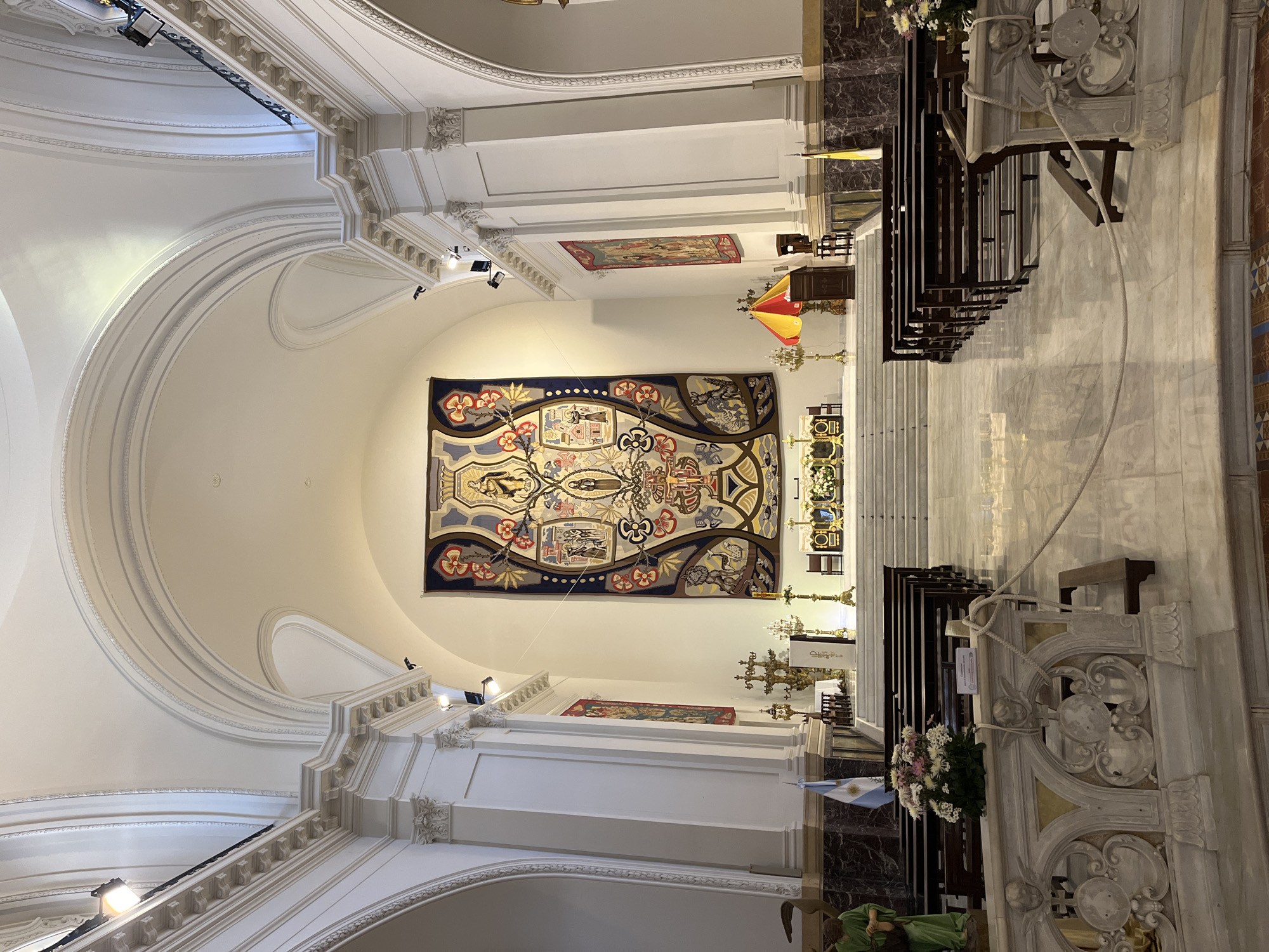 The media file shows the grand interior of a church or cathedral, viewed from a slightly elevated position looking down a side aisle towards the altar area. The setting is likely in Buenos Aires, Argentina, as indicated by a prominent Argentinian flag.
The scene is dominated by classical architectural elements: high, vaulted white ceilings, ornate cornices, pilasters, and arches along the left side. The walls are also painted white, creating a bright and expansive feel.
On the right side of the image, rows of dark wooden pews are neatly arranged alongside a wide, light-colored marble aisle. In the immediate foreground on the right, an intricately carved white marble or stone balustrade or partition features elaborate Baroque-style relief work, with a rope tied to it. A fresh floral arrangement of white and pink flowers is placed nearby, accompanied by a draped Argentinian flag. A green-robed statue of a reclining figure is partially visible at the bottom right.
On the left wall, a large, colorful tapestry hangs prominently, depicting religious or symbolic figures and motifs in a detailed, stylized manner. Below it, a smaller, reddish tapestry is also visible. The altar area, located towards the back and center-left, is adorned with ornate golden candlesticks and crosses. Another flag, possibly a Vatican flag, is seen further back near the pews.
The space is illuminated by artificial spotlights directed at the tapestries and architectural details, suggesting either evening or a dimly lit interior. There is no discernible activity taking place, and the church appears largely empty. A small, white sign is visible on the ornate balustrade on the right, but any text on it is illegible. The overall impression is one of grandeur, history, and religious solemnity.