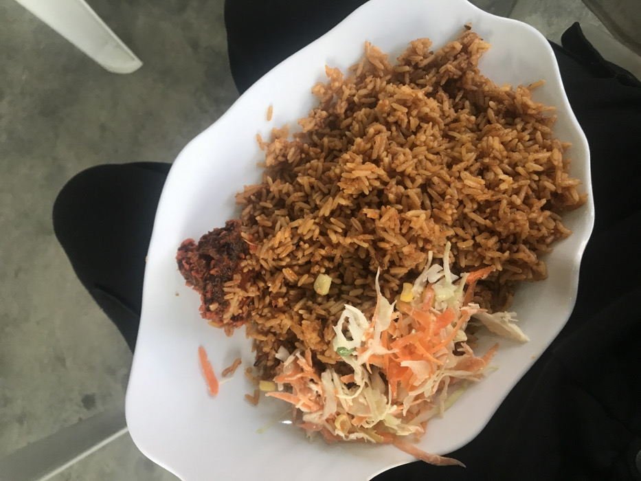 A white plate is filled with cooked rice, which is brown. Next to the rice is a piece of fried chicken, which has a darker brown color. A side of coleslaw is also present on the plate, consisting of shredded cabbage and carrots. The plate is set on a surface that includes black fabric.