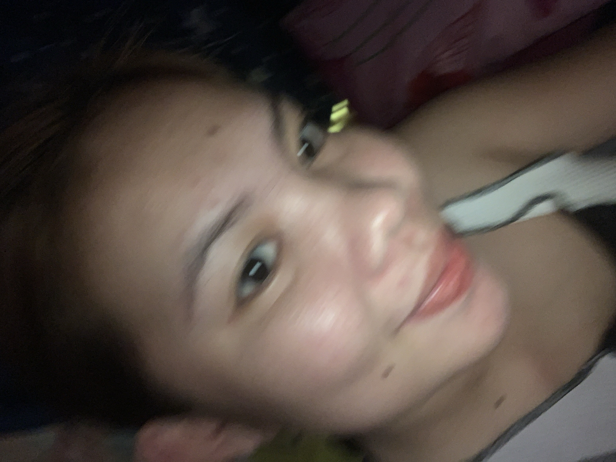 A selfie is taken from a close-up, low-angle perspective of a young woman with light brown skin and dark brown hair. Her face is slightly blurred, but she appears to be smiling with her mouth slightly open, revealing a glimpse of her lips. She is wearing a dark-colored top with white trim. The background is dark and appears to be indoors, with some blurry hints of bedding or fabric. There is no discernible indication of the city of Kiblawan, Philippines.