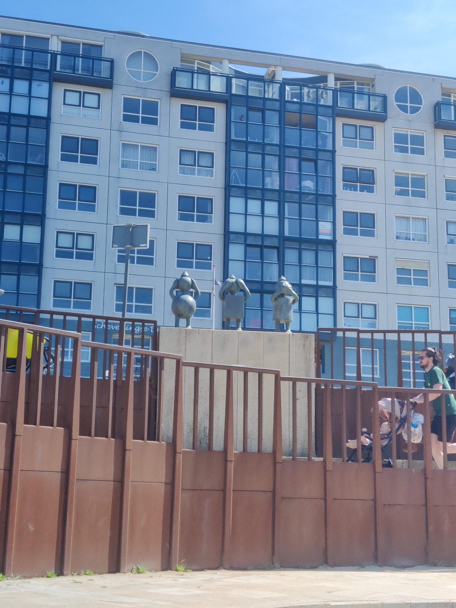 In The Hague, Netherlands, an apartment building with a grey facade and blue-tinted windows stands under a clear blue sky. The building features multiple floors of windows and balconies. In the foreground, a rusty brown metal fence runs horizontally across the image, with vertical bars. On top of a concrete structure behind the fence, three bronze statues of figures with round heads and arms akimbo are positioned. To the right of the statues, a man wearing a green t-shirt and black shorts walks while pushing a stroller. The stroller appears to have a baby inside.