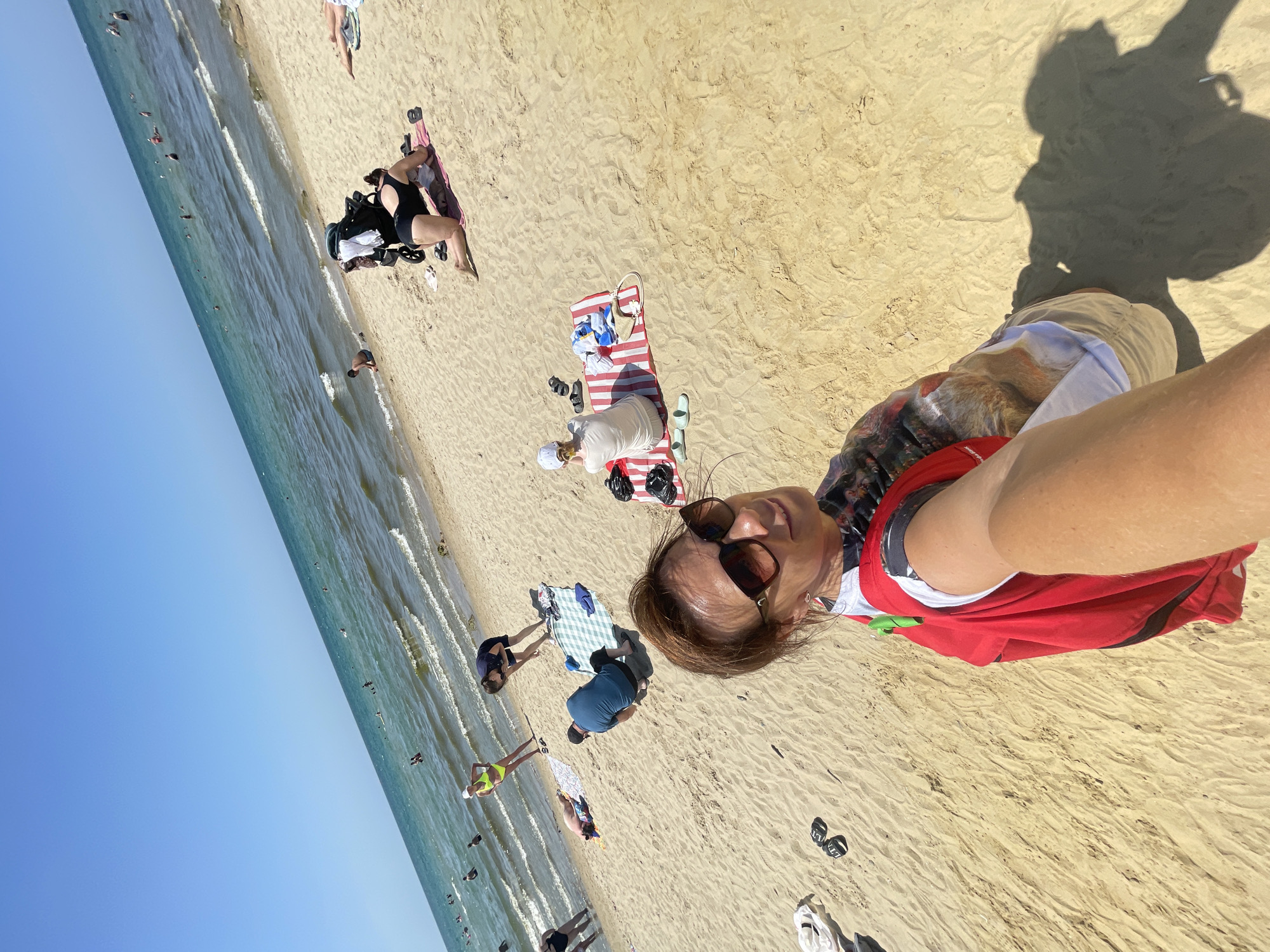 The image depicts a sunny day at a beach, possibly in Anapa, Russia, with several people engaged in various activities. In the foreground, a woman in sunglasses and a red cover-up is taking a selfie. In the middle ground, several individuals are spread out on the sand. Some are sitting on towels, one person is in a black swimsuit on their hands and knees, and another is seen near the water's edge. In the background, the sea is calm with small waves, and numerous people are swimming or wading in the water. The beach itself is sandy, and the sky is clear blue.