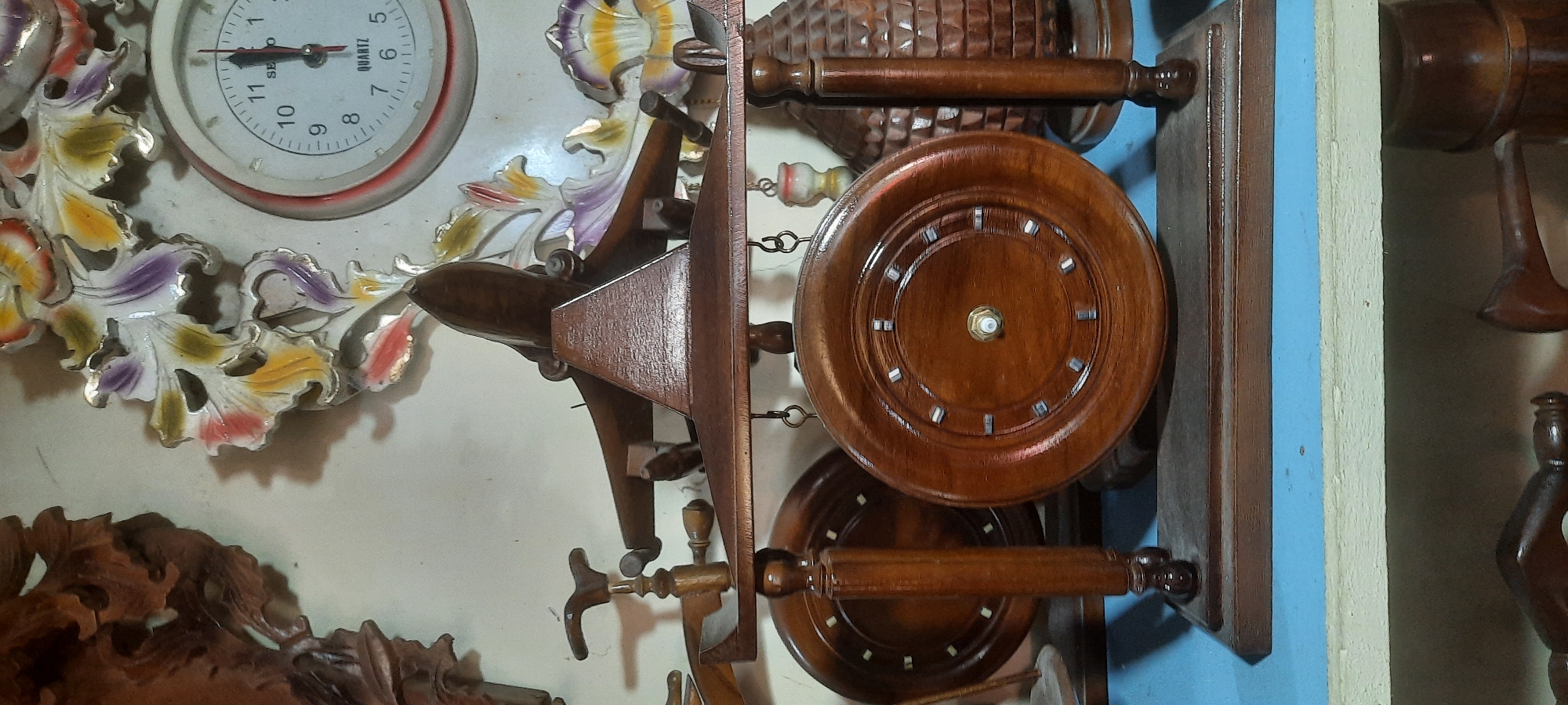 The image shows a close-up view of a wooden clock with an airplane on top, displayed in what appears to be a shop or home interior in Blitar, Indonesia. The clock itself is ornate, featuring carved wooden elements and a face with Roman numerals. The airplane is also made of wood and is positioned to look like it's in flight. In the background, there are other decorative items, including what looks like a colorful, floral-themed clock and a carved wooden panel. The lighting suggests an indoor setting, possibly a shelf or display area.
