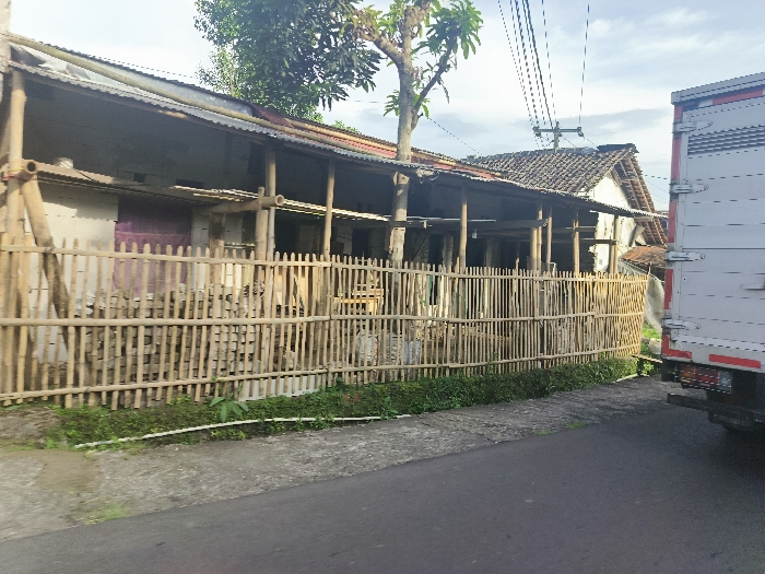 The image shows a partially obscured view of a residential building in Cikalong, Indonesia, located at coordinates -6.732 latitude and 107.441 longitude. The building has a corrugated metal roof and wooden support beams. The building is enclosed by a bamboo fence. A white truck is parked on the asphalt road outside the fence. The truck is facing away from the viewer. The road appears to be empty, with no people visible in the scene. There are power lines overhead, and a green tree with a few branches is visible in the background. The scene is quiet and appears to be taken in a rural area. The building is in a state of partial construction or renovation, with exposed wood and beams.