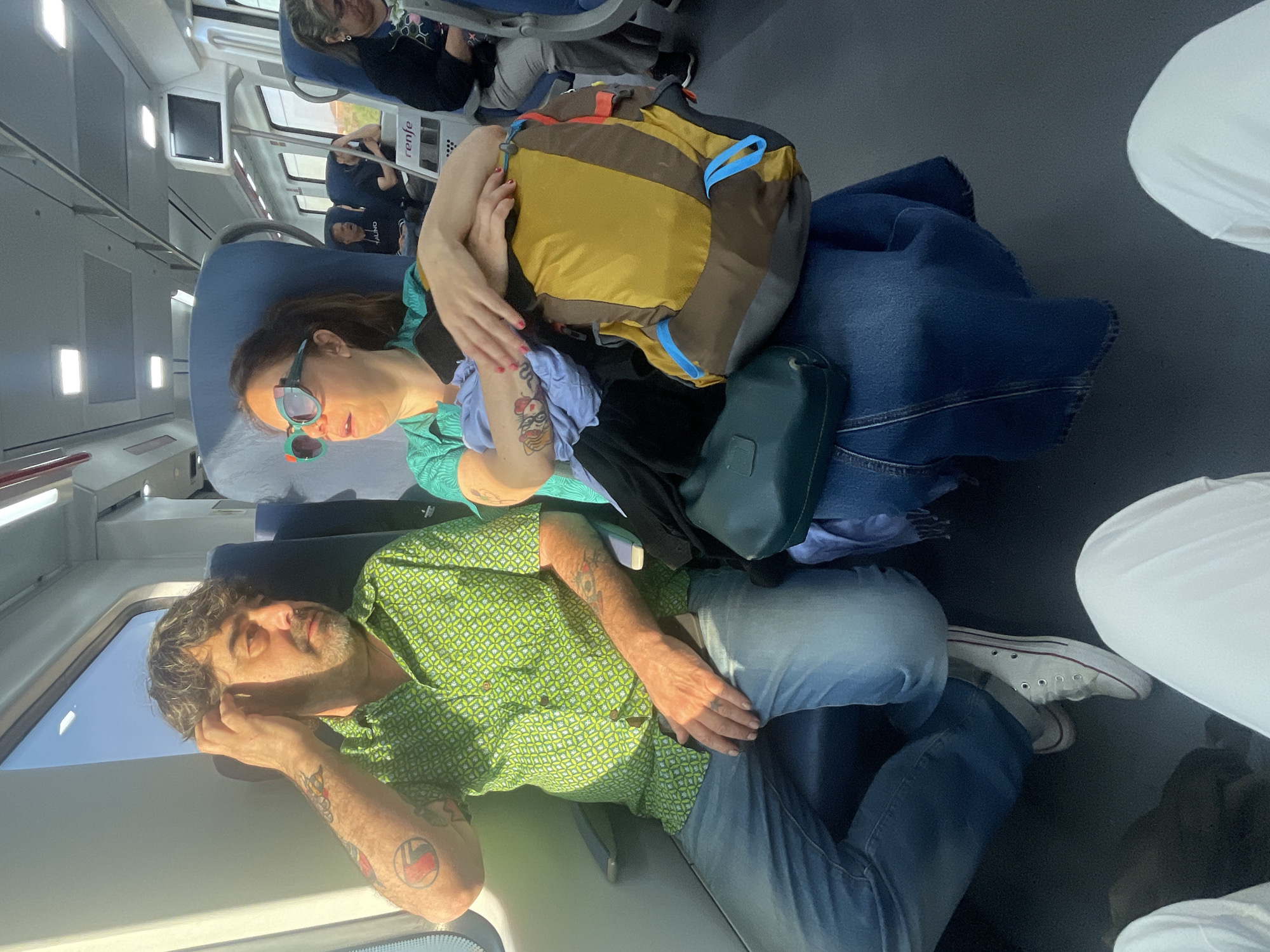 Two individuals are seen on a train. A man with curly hair and tattoos lies near the window wearing a green patterned shirt and jeans. A woman wearing sunglasses and a green top rests with her arms around a yellow and brown backpack. Behind them, other passengers sit. The train's interior includes blue seats, windows, and overhead compartments. The image appears to have been taken on a train, not in Les Sènies.