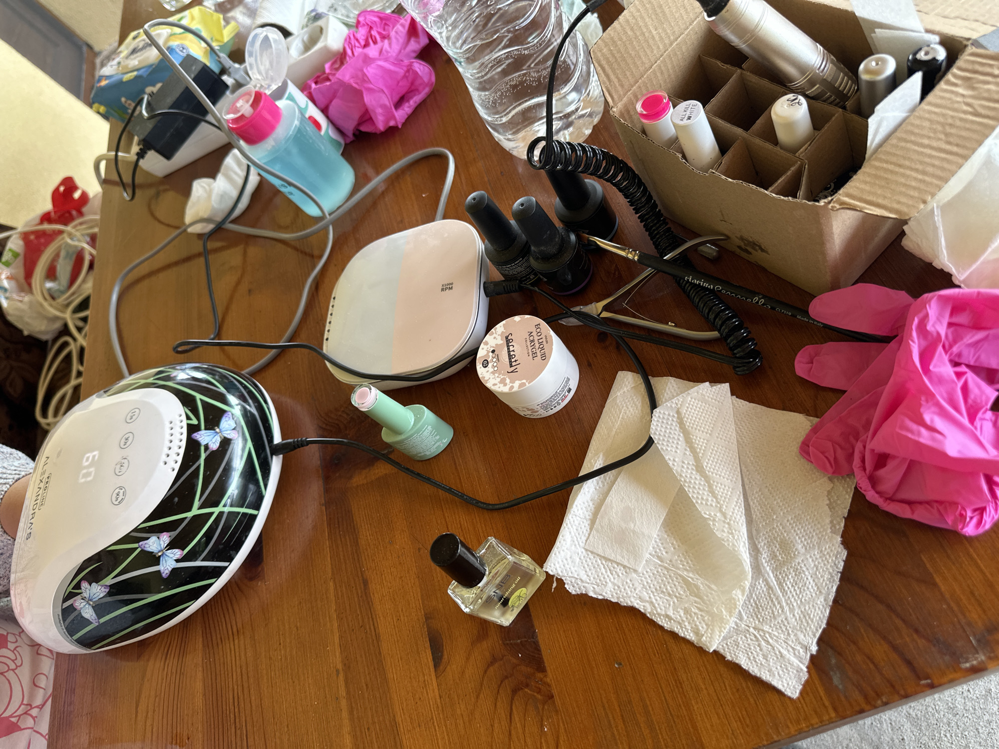 The image shows a wooden table surface cluttered with various nail care items. A nail lamp with butterfly decorations sits on the left side of the frame. Nearby are bottles of nail polish, including one clear bottle labeled "SNB nourishing oil". There's also a container labeled "ECO LIQUID ACRYGEL COLLECTION" from "secretly". A cardboard box holding nail polish bottles is on the right. Several pink gloves, white paper towels, and a water bottle are scattered around the table. A brush labeled "clarina Connello CL7319 Made in Italy" is also visible. The location in the city of Lehchevo, Bulgaria is not discernible.
