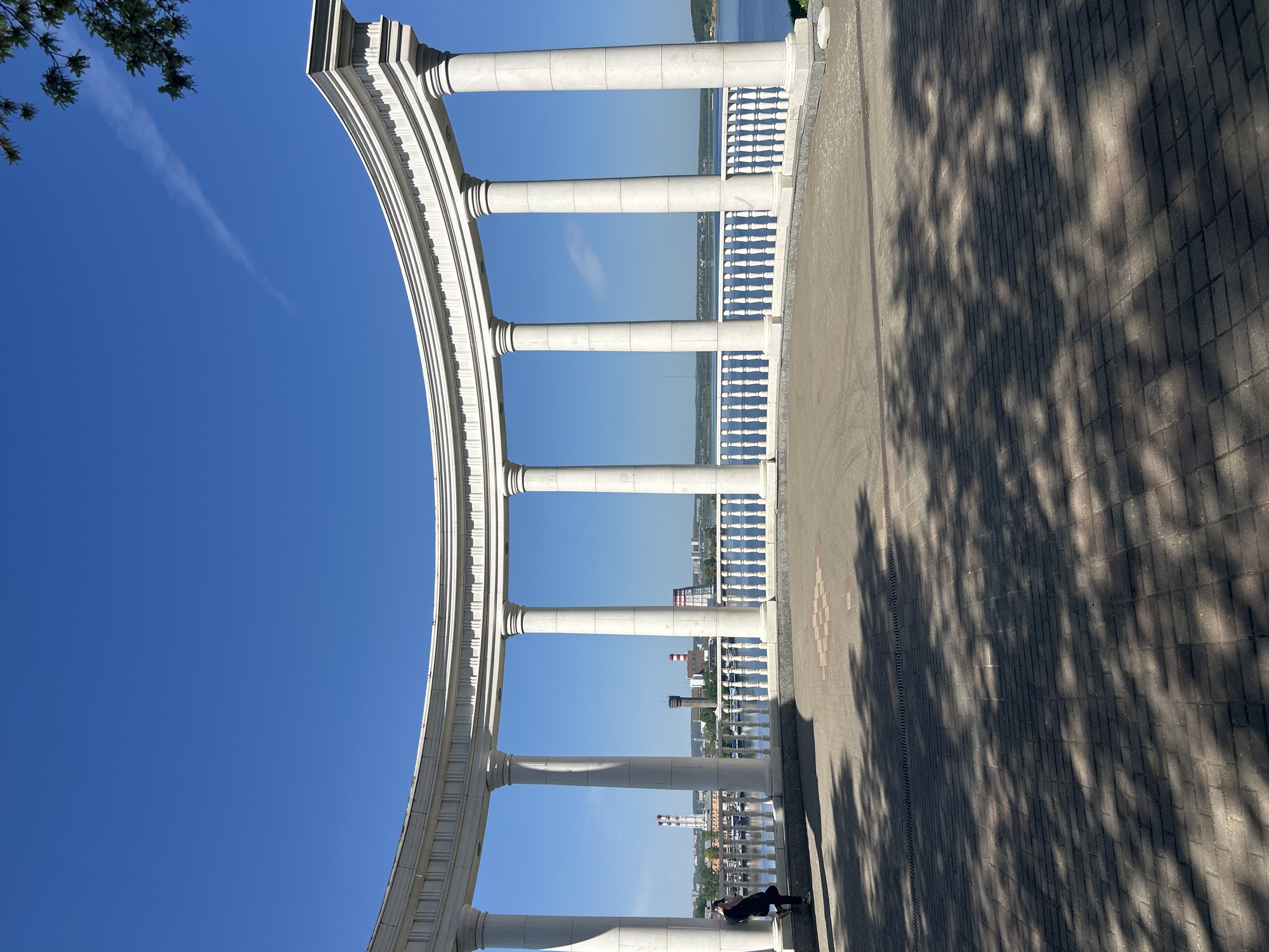 The image depicts a colonnade overlooking a body of water, possibly a river, with buildings visible in the distance. The colonnade is made of white stone and has a curved shape. A person is lying on the base of one of the columns. The ground is paved with bricks. Tree shadows are cast on the ground. The sky is blue and clear. The location is likely in Izhevsk, Russia, given the industrial architecture and river setting consistent with the city.