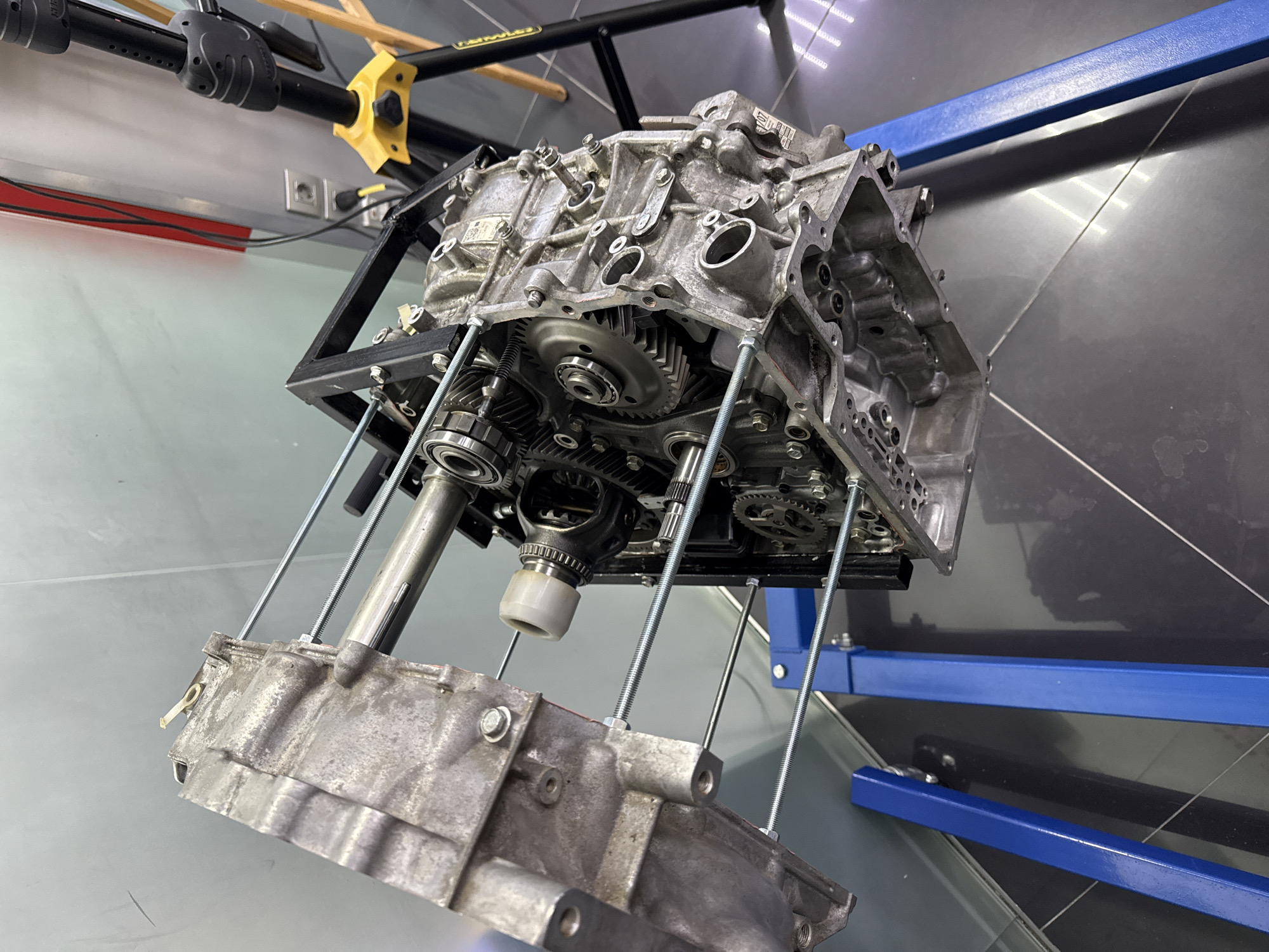 The image shows a partially disassembled engine block, likely a transmission, suspended in a black metal frame with four long threaded rods. The engine components, appearing metallic gray, consist of gears, shafts, and the engine casing. The lower part of the engine casing is partially detached. The supporting frame rests on a blue metal stand. The background features a tiled floor with a dark gray surface, and in the upper area, there is equipment, a yellow support, and an electrical outlet. The setting appears to be a workshop or garage, as suggested by the maintenance-related equipment and tools. There is no indication of Perm, Russia in the image.