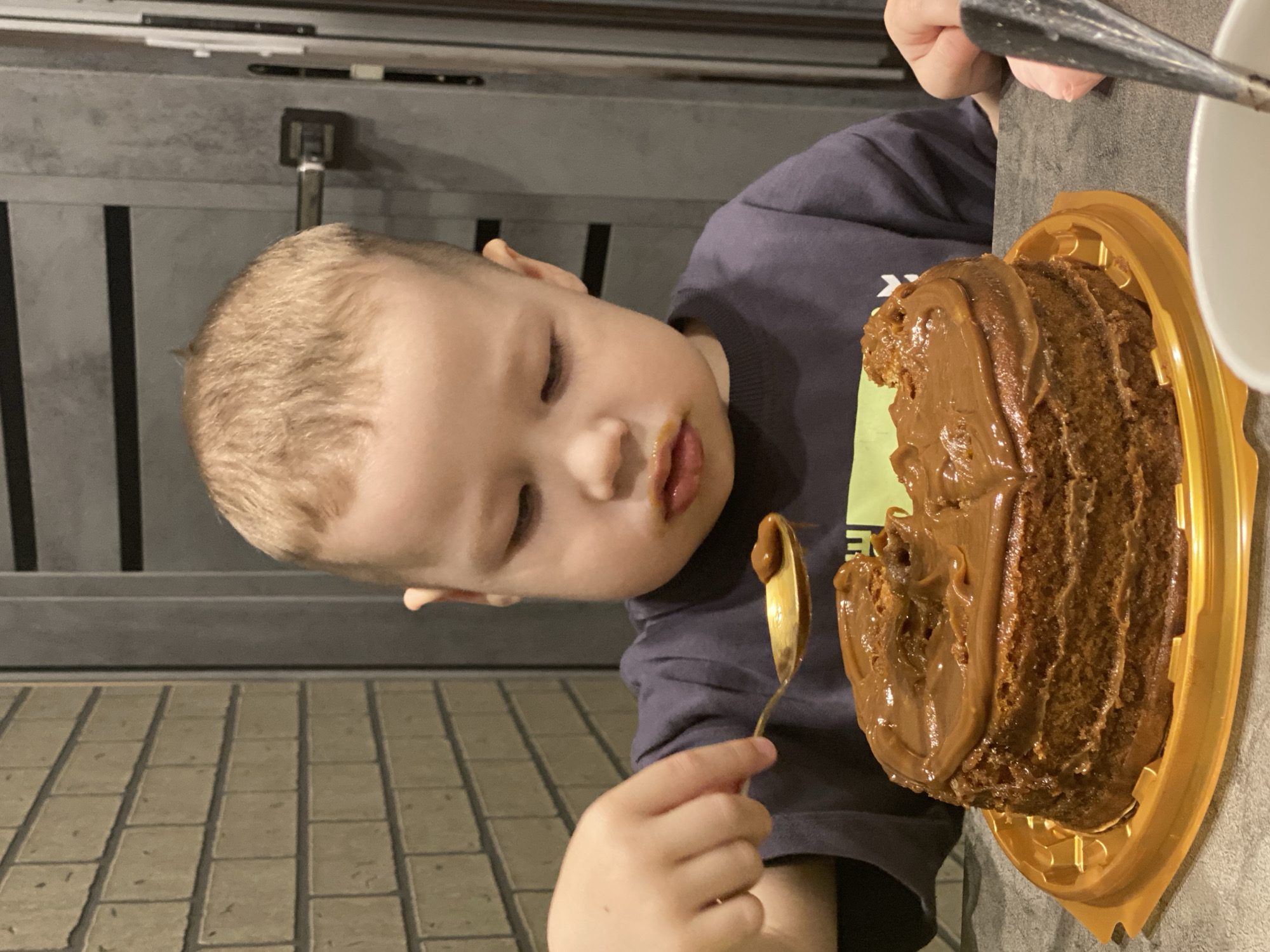 A young boy is captured indoors, engrossed in eating a generously frosted cake with a spoon. He has light brown hair and is wearing a dark t-shirt with a subtle light green design partially visible. His eyes are partially closed in concentration as he scoops a spoonful of light brown frosting from the cake, with some frosting also visible around his mouth. He holds a golden-colored spoon in his right hand.
The cake is round, dark brown, and appears to be chocolate or caramel flavored, covered in a thick, shiny, light brown frosting. It rests in a golden, ridged plastic container. A portion of the cake and frosting has already been eaten.
The background shows an indoor setting with dark grey, vertically paneled walls or a door in the upper part of the image, and a light-colored, square-tiled floor in the lower part. The lighting is soft and even, typical of an indoor environment. The child appears focused and content while enjoying his dessert. No legible text is visible. The location is Sochi, Russia.