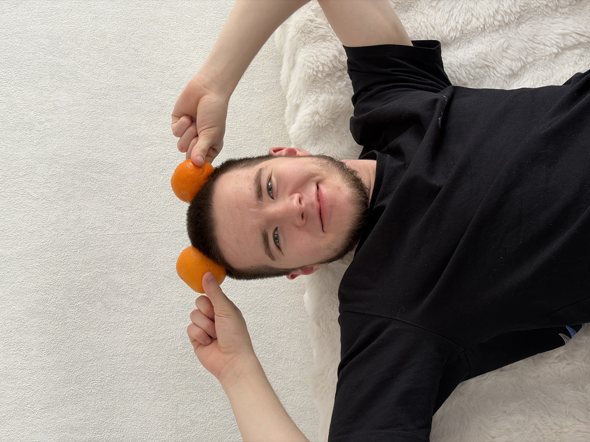 A man is lying on his back on a fluffy white surface, possibly a rug or a blanket. He is wearing a black T-shirt. He is holding two tangerines, one in each hand, and positioning them on either side of his head, seemingly balancing them. The background is a plain white wall. The location appears to be indoors.