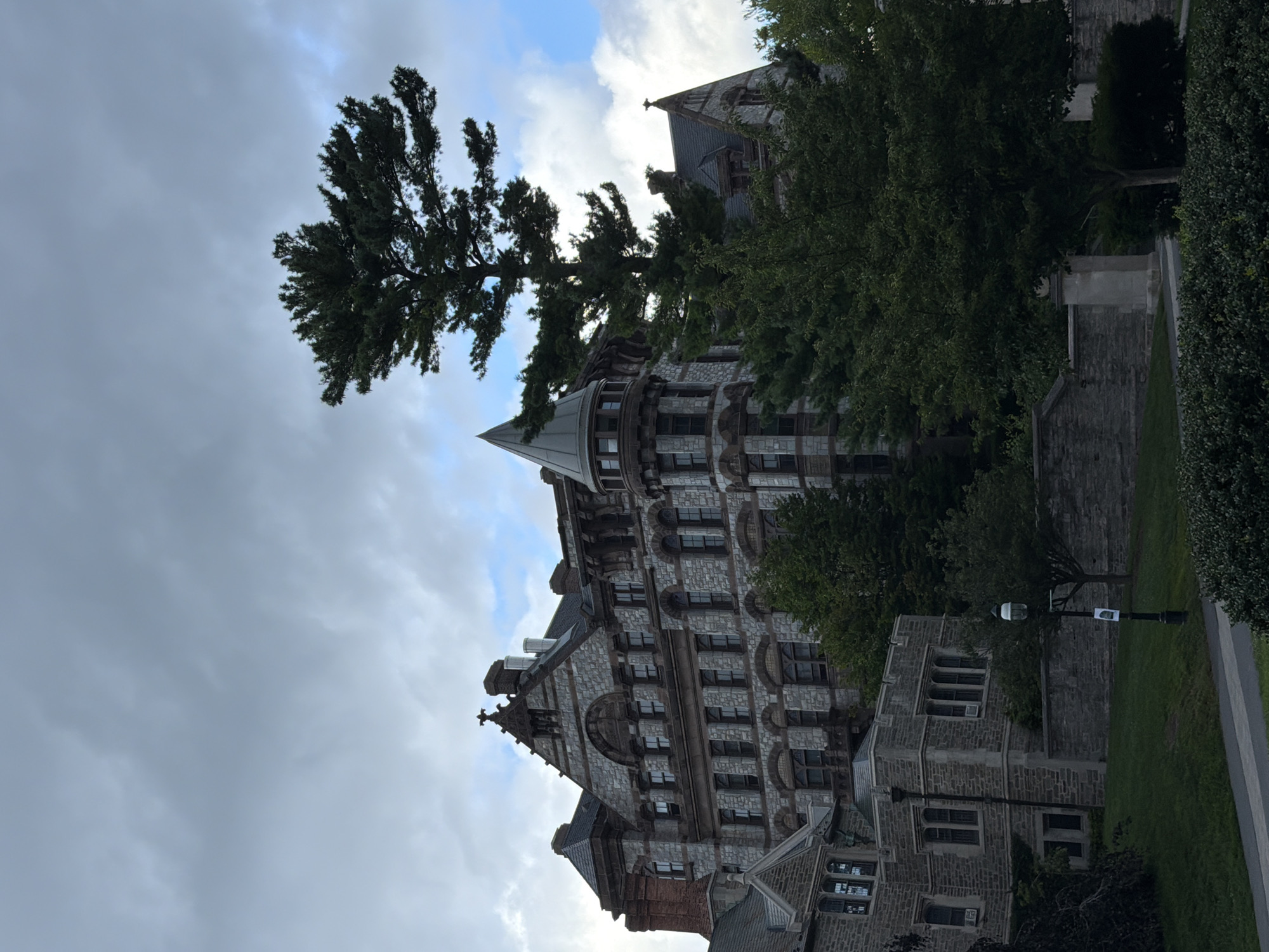 The image shows the exterior of a large, ornate stone building with a gothic architectural style, likely a university building in Princeton, New Jersey, United States of America. The building features multiple levels, arched windows, decorative stonework, and several chimneys. A prominent, tall coniferous tree stands in front of the building, partially obscuring it. To the left of the main building, a smaller, similarly constructed stone building is visible. The scene is set on a grassy area with a paved walkway in the foreground. A lamppost is also present on the walkway. The sky is overcast with some patches of blue sky visible. The overall impression is one of historical architecture and a serene, academic environment.