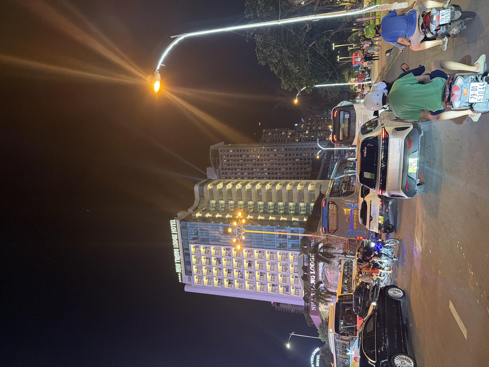 This detailed image captures a vibrant night scene in Nha Trang Ward, Vietnam, showcasing a multi-lane urban street with heavy vehicle traffic and illuminated buildings.
In the foreground, a dark asphalt road dominates, marked with white solid and dashed lines. Numerous vehicles are present, indicating active traffic. Prominently, two individuals in the immediate foreground are riding scooters away from the camera: one wearing a green shirt and white helmet on a scooter with license plate 79-01 530.44, and another in a blue shirt and green helmet on a scooter with license plate 79-N1 375.57. A third scooter, positioned further ahead in the center of the road, carries three occupants: a driver and two passengers, one wearing a red shirt and the other a dark shirt. To the left, a black sedan is partially visible. Behind the central scooter, a white VinFast SUV with "XE TAXI" signage and license plate 79H 174.78 is stopped.
The mid-ground features several large tour buses. A blue bus with "UNIVERSE" branding and license plate 50F-017.84 is prominent on the right. Yellow and white buses are visible further left.
The background is dominated by towering, illuminated buildings against a dark night sky. The most prominent structure is the "NHA TRANG LODGE HOTEL," a multi-story building with a white and blue glass facade. Its numerous balconies are lit with warm yellow light, and a green neon sign spells out its name at the top. Below the main sign, "Nha Trang Lodge" is visible on a lower section. To the right of this hotel, other high-rise buildings, also illuminated, contribute to the urban skyline. A partial view of another building on the far left reveals "EN VIEN DONG HOTEL."
Multiple streetlights punctuate the scene. A bright yellow-orange streetlight on the right emits strong light rays, and another decorative streetlight with multiple arms is centrally located. Other streetlights, some with white light, line the road and a tree-lined area on the far right. Palm trees are visible near the base of the Nha Trang Lodge Hotel. On the far right, a sidewalk area is populated by several pedestrians strolling under the ambient light. The overall atmosphere is bustling and brightly lit, indicative of an active urban center at night.