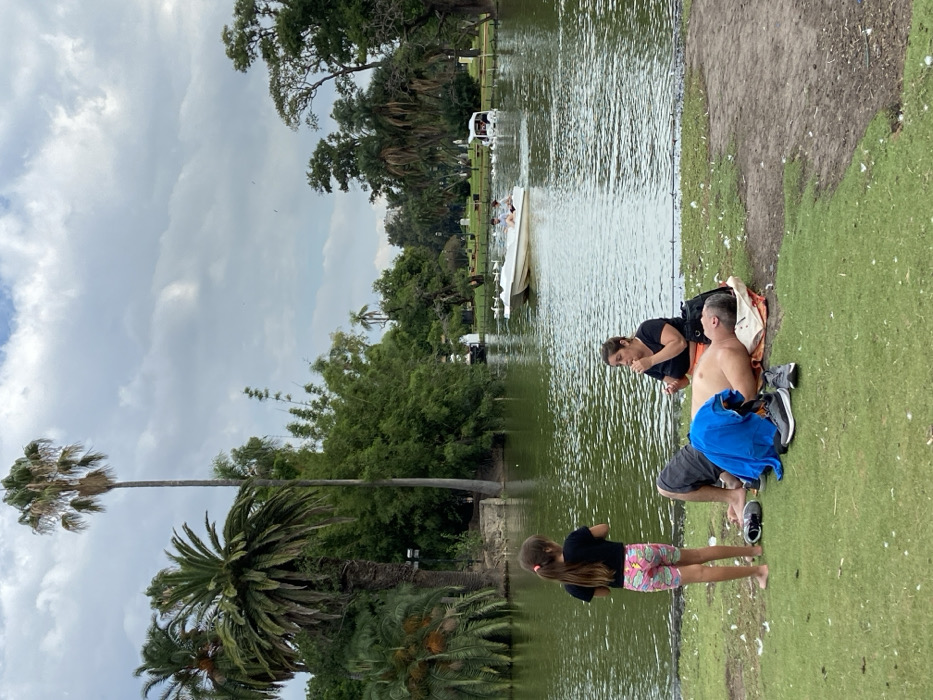 A family sits beside a pond. A man is shirtless and beside a woman with a black shirt. A girl stands on the grass wearing a black shirt and patterned shorts. Several boats are in the pond. Lush trees and plants surround the pond and a tall palm tree is also visible. The sky is cloudy.