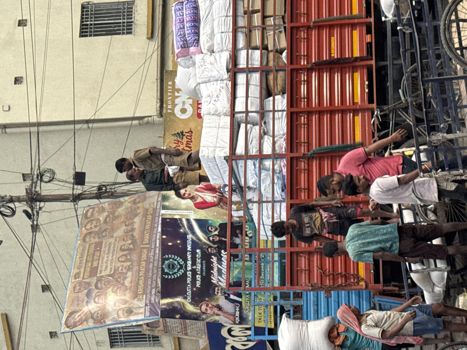 A busy street scene features multiple advertisements and people. A large red truck is loaded with white sacks, possibly goods for sale. Several men are working or riding on the truck. Underneath the truck, a banner reads "SRI KRISHNA PRATAP SINGH SHAKTI PRATAP SINGH, CALCUTTA POLICE SERGEANTS INSTITUTE & POLICE ATHLETIC CLUB CELEBRATE Midnight Madness." Above the truck, another sign includes "FRONTIER" and "Christmas." Utility poles with numerous wires are visible against a building.