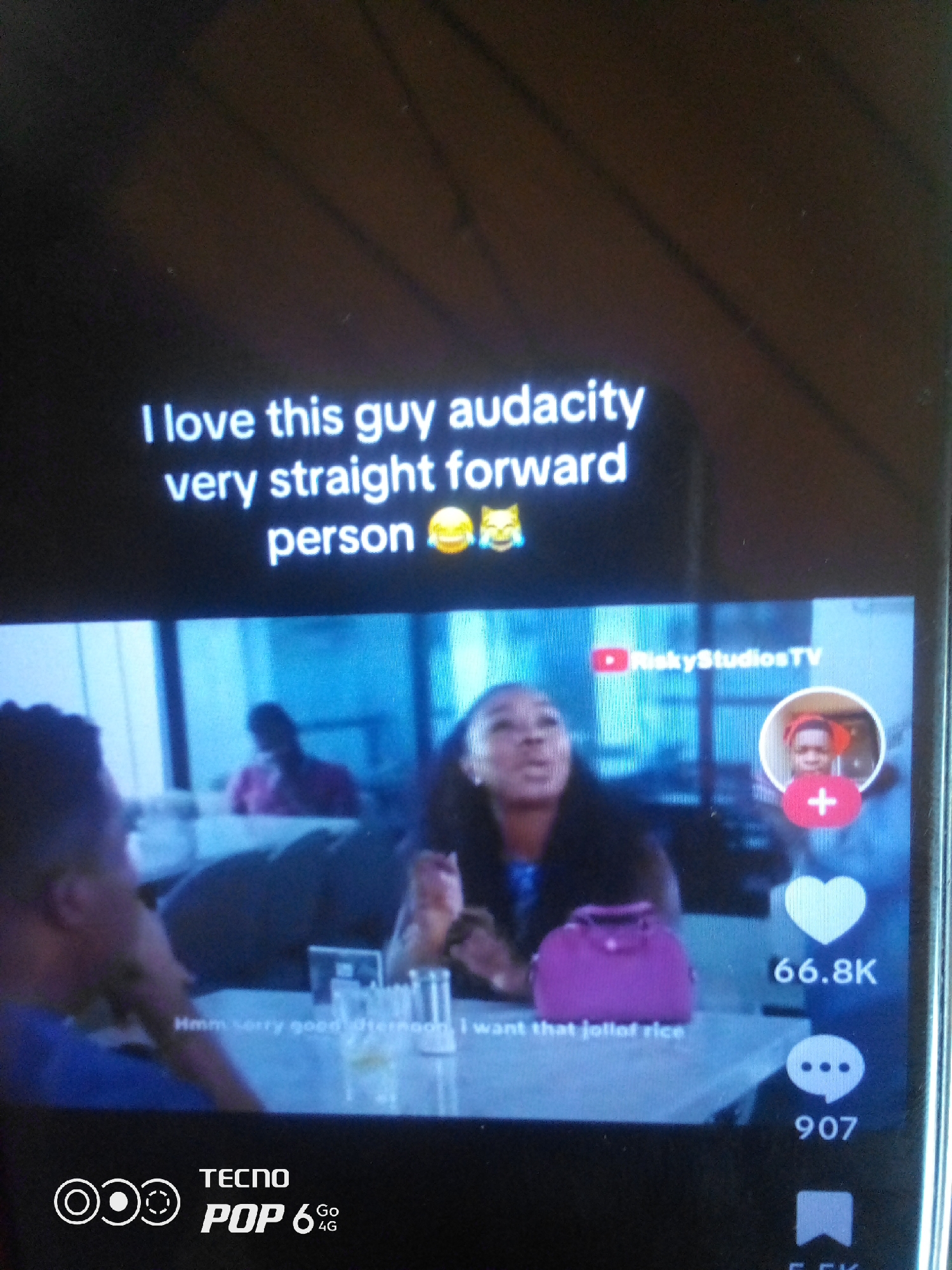 The image is a screenshot of a mobile phone displaying a video from a social media platform, likely TikTok or a similar app. White text overlaid on the video reads, "I love this guy audacity very straight forward person ๐" with two laughing emojis. Below this text, a video plays showing several individuals in what appears to be a restaurant or indoor social setting. In the foreground, the faces of at least two people are partially visible. In the middle ground, a woman is speaking, and to her right, a circular profile picture with a plus sign indicates a user's channel, displaying the name "RiskyStudiosTV" with a YouTube logo. On the right side of the screen, social media interaction icons are visible: a heart with the number "66.8K" next to it, and a speech bubble icon with "907" below it. The bottom of the screen displays the TECNO POP 6 4G logo. The location is not specified in the image.