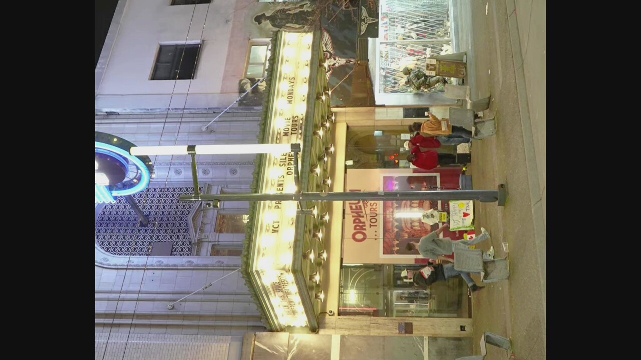 The image depicts the Orpheum Theatre in Vancouver, Canada, at night. A marquee with illuminated lettering announces "VGT PRESENTS SILENT MOVIE TOURS" and "ORPHEUM TOURS MONDAYS". Several individuals are visible in front of the theatre. Two men are walking past a row of metal chairs in the foreground. Further back, a group of people stands near the entrance, some wearing winter clothing. A sign for "ORPHEUM TOURS" is posted on the left side of the entrance. To the right, a large mural featuring a winged figure is partially visible. A metal gate encloses an area to the far right. The facade of the theatre is ornate, with decorative stonework and a large arched window above the marquee. Streetlights and the theatre's lights illuminate the scene.