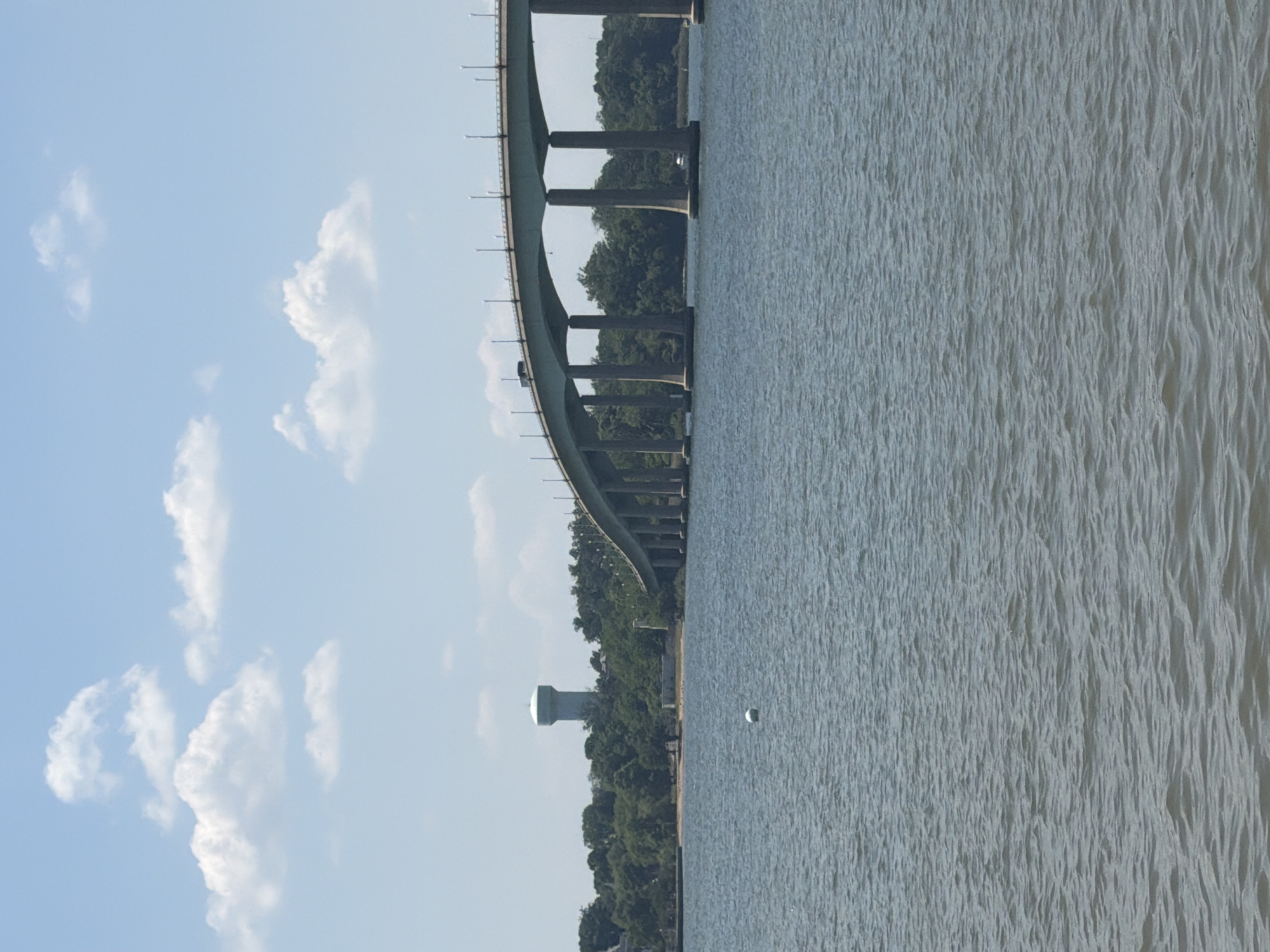 The image captures a view of the Severn River, Maryland, adjacent to the United States Naval Academy. A section of the Severn River Bridge is visible on the left side of the frame, arching over the river with its support pillars prominent. The river's surface is rippled, indicating a slight breeze. Adjacent to the bridge's base, trees line the riverbank, with a water tower visible among them. The sky overhead is light blue with scattered cumulus clouds.