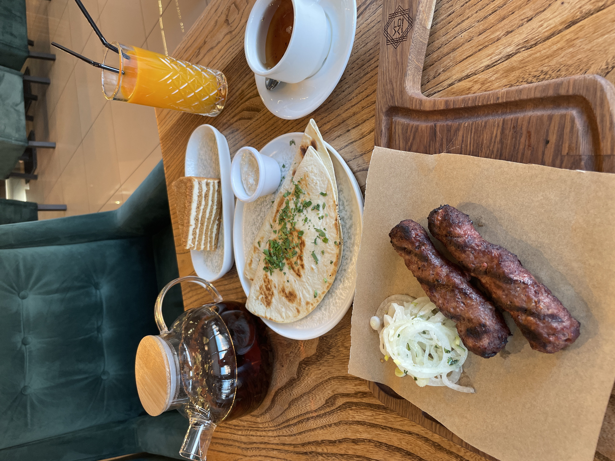 A high-angle, close-up shot shows a table setting in what appears to be a cafe or restaurant. The setting includes two grilled meat skewers, sliced onions, a piece of layered cake, a glass of orange juice with two straws, a cup of tea with a saucer and spoon, a small bowl of white sauce, and a glass teapot filled with dark tea. The table is made of wood with a prominent grain. In the background, a plush green velvet chair is visible, along with glimpses of other chairs and tiled flooring, suggesting an indoor dining environment. A wooden serving board with carved detailing is also part of the arrangement. The overall impression is of a meal being served.