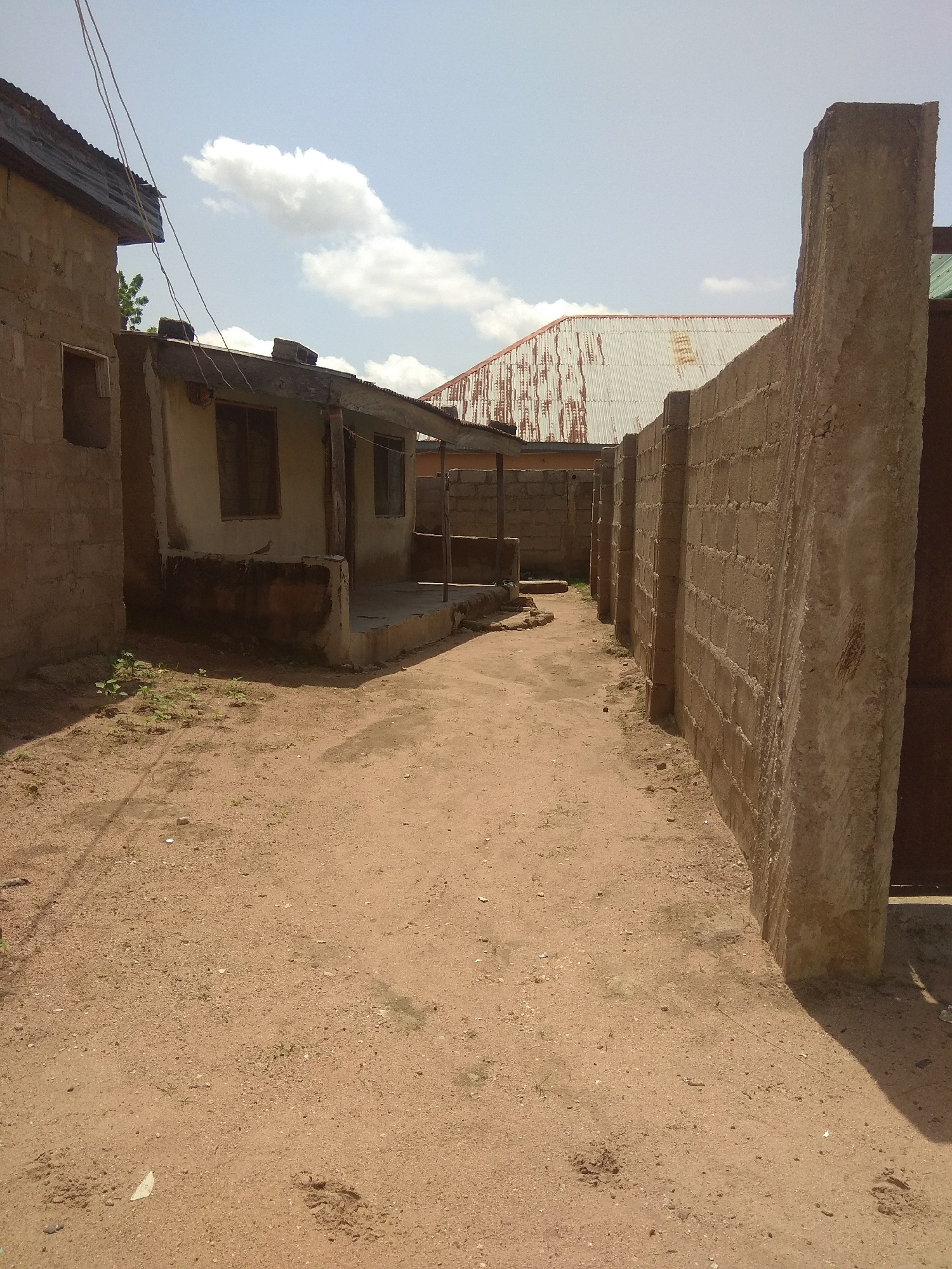 The image depicts a dusty alleyway between two buildings in the city of Jalingo, Nigeria. On the left, a light brown, unfinished concrete block building with a corrugated metal roof is partially visible, featuring an empty window frame and a dark, recessed doorway. To the right, a tall, rough concrete wall encloses the alley, with a weathered, corrugated metal roof visible in the background, attached to a building with arched sections. The ground is covered in reddish-brown sand and small pebbles, with a few scattered green plants and some indentations suggesting footprints. A covered walkway, supported by wooden posts, extends from the building on the left into the alley. The sky above is partly cloudy with a pale blue hue.