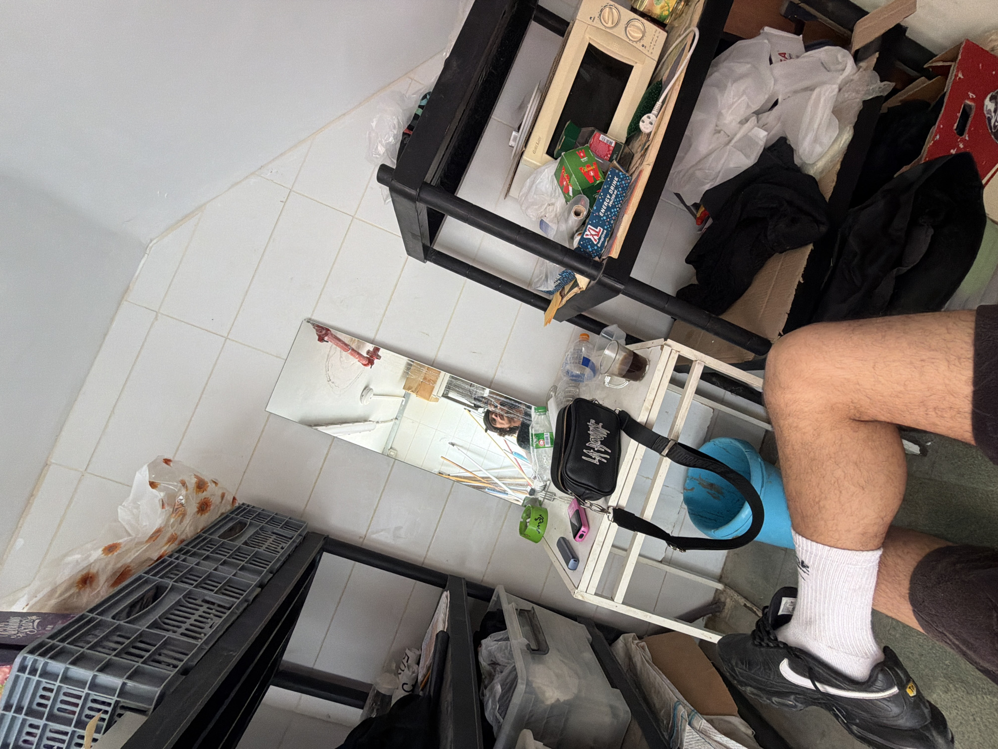 The image presents an indoor scene captured from a low, tilted angle, looking across a cluttered room. The floor is laid with light-colored square tiles, and the walls are similarly light.
In the foreground on the right, a person's legs are visible, wearing dark shorts, white socks with a small black logo, and black Nike sneakers featuring a white swoosh and "Tn" branding. The person's reflection, showing a head with dark curly hair, is visible in a tall, possibly fragmented, mirror positioned on the left side of the frame.
The room is filled with numerous objects. To the immediate left of the person's legs, a white shelving unit or table holds several items, including a clear plastic water bottle, a bottle with a dark liquid, a pink mobile phone, a small black vape device, and a green wristband or bracelet. A black cross-body bag, bearing the white text "LA BOUGE", hangs from this unit. A bright blue bucket or container is positioned near the person's feet.
In the background, a black metal shelving unit is heavily loaded with various items. On its upper shelf, a small, beige microwave oven, partially labeled "Gold...Melt...", is visible. Other shelves contain boxes, including one prominently displaying "ENERGY DRINK XL" and another labeled "Classic". The unit is also piled with white plastic bags, folded dark clothing, cardboard, and other miscellaneous items. A gray plastic crate is visible on the far left.
The scene appears brightly lit, suggesting it is daytime. The environment is notably untidy, resembling a storage area, utility room, or a very messy domestic space. The activity captured is the person taking a self-portrait or mirror selfie. The location context provided indicates the scene is in Hof HaCarmel Regional Council, Israel.