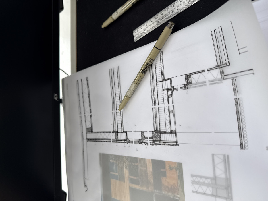 The scene depicts a desk setup located in Newcastle upon Tyne, United Kingdom, centered on architectural documentation. A white sheet of paper, displaying technical architectural drawings, occupies the midground. The primary drawing is a detailed cross-section illustrating building wall and floor construction elements, rendered in black lines and hatching. To the left, a blurred photographic image of a modern building with wooden cladding is visible underneath or alongside the main drawing.
A beige-bodied technical drawing pen, identified as a Pigma Micron 005, rests horizontally across the architectural drawing. To the right of the paper, on a dark, flat surface, a second identical pen is partially visible along with a metallic ruler, approximately 15 cm (6 inches) in length, showing both metric and imperial markings. The background features the bottom bezel of a black computer monitor, with the "ADX" brand logo visible. The monitor screen is dark. No individuals are present. The arrangement suggests an active architectural design or study environment.