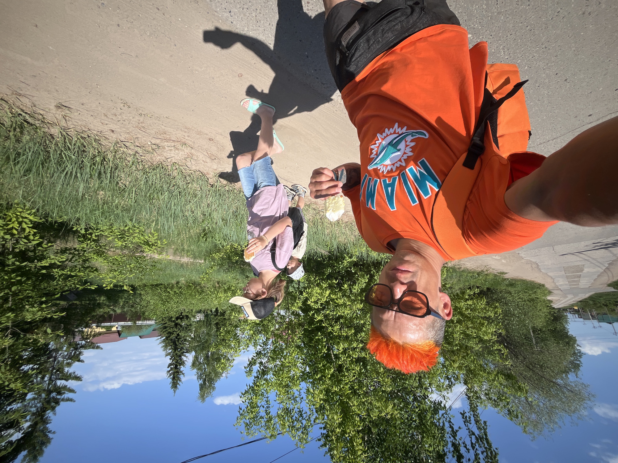 A man and a child are walking on a dirt road in Chaikovsky, Russia. The man, positioned closer to the camera, is wearing an orange Miami Dolphins t-shirt, black shorts, and orange-tinted sunglasses. His hair is dyed orange. He is holding an ice cream cone. The child, walking ahead of the man, is wearing a pink shirt, denim shorts, and teal sandals. They are also holding what appears to be an ice cream cone. The road is lined with grass and trees. The sky is blue and clear. The houses, trees, and sky are partially visible in the background.