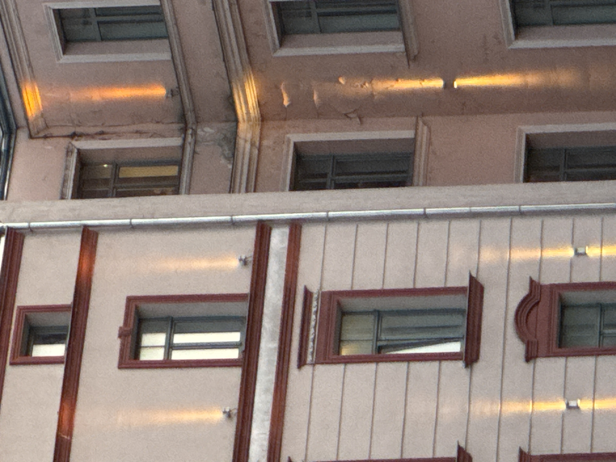 The image shows a close-up view of the facade of two adjacent buildings. The building on the left has a textured, light-colored wall with several windows framed by dark red trim. Some of these windows are rectangular, while one in the upper section is a narrow, tall rectangle. The building on the right has a pinkish-tan colored wall, also with windows. The windows on this building are also framed by trim, appearing white in the upper section and dark red in the lower section. Both buildings feature exterior lighting that casts vertical beams of orange light onto the walls, highlighting architectural details. A vertical metal pipe or conduit runs along the seam between the two buildings. The scene appears to be an urban environment, likely a street in a historic district, given the architectural style. The image quality is good, with clear details of the building materials and features. The lighting suggests it might be evening or early morning, or that the lights are intentionally illuminated. The specific location within Riga, Latvia is not visually indicated.