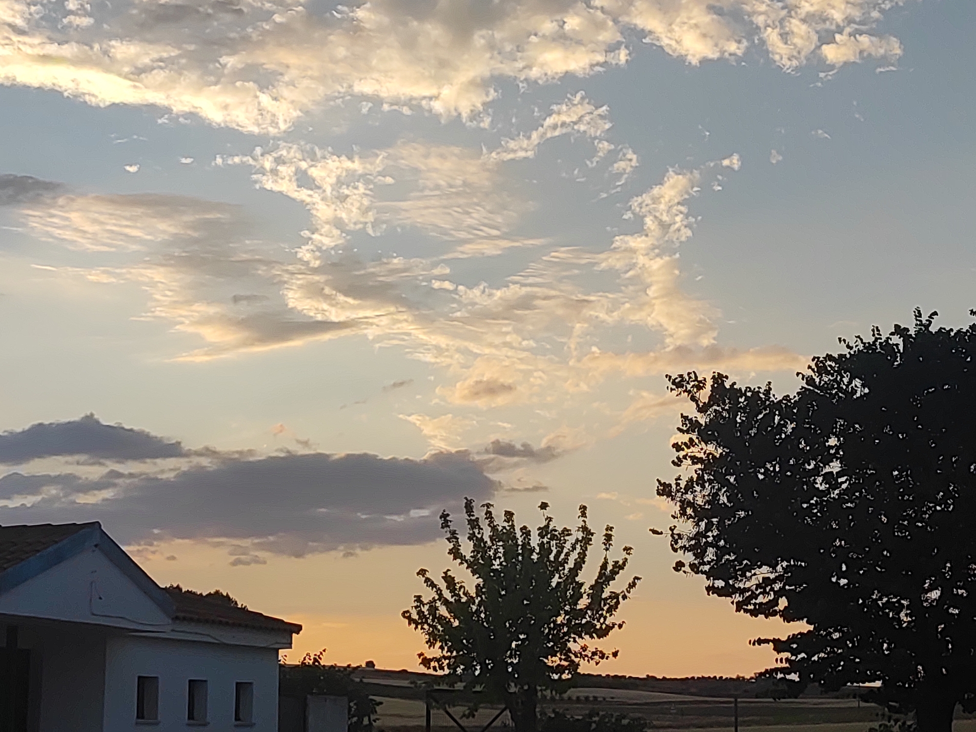 The image captures a rural landscape at dusk in Villarejo de Fuentes, Spain. The sky is a gradient of soft orange and pale blue, filled with wispy, illuminated clouds. In the foreground, on the left, is the side of a white building with a blue-trimmed roof and three small, rectangular windows. To the right of the building, a sparsely leaved tree stands silhouetted against the sky. Further to the right, a large, dark tree dominates the frame, its dense foliage obscuring much of the horizon. The horizon itself is low and indistinct, with subtle hints of cultivated land and distant structures. The overall atmosphere is serene and tranquil, indicative of the end of the day.