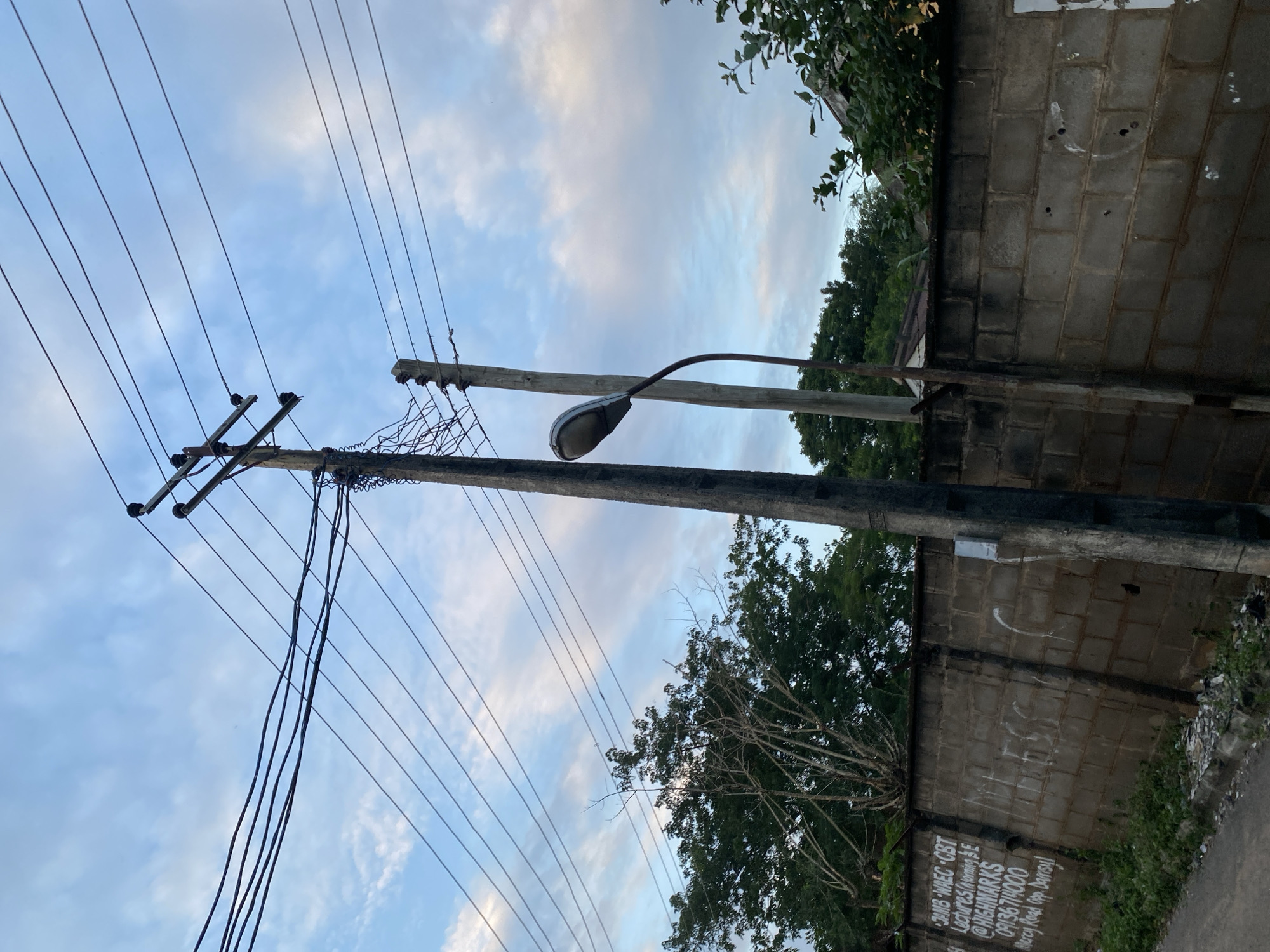 The image shows a street scene in Abeokuta, Nigeria, with a focus on utility poles and electrical wires against a cloudy sky. The foreground features a concrete block wall with some graffiti, including what appears to be contact information. A street lamp is visible, attached to one of the poles. Trees and foliage are present in the background, suggesting an urban or semi-urban environment. The overall impression is one of a typical roadside in the city.